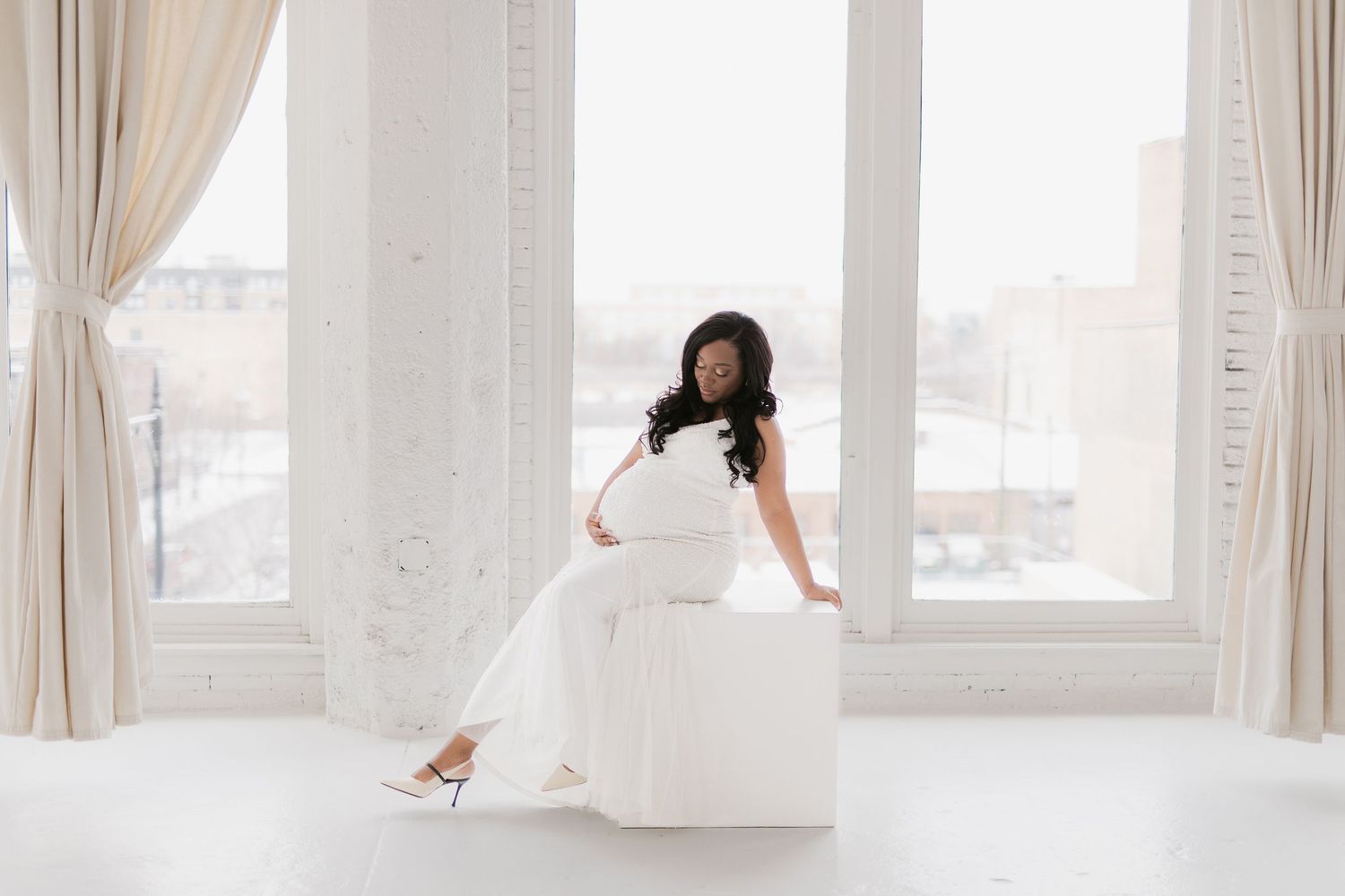 Pregnant woman in white dress sitting by large window in bright studio, maternity photography session.