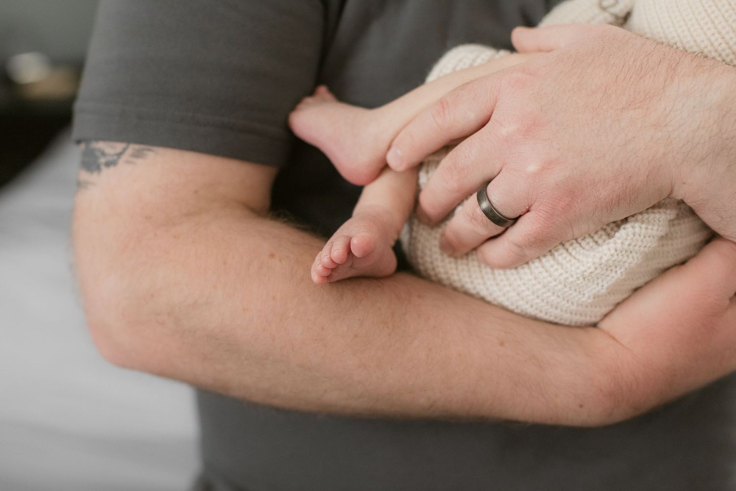 Close-up of hands cradling a newborn baby wrapped in white fabric with a wedding ring visible.