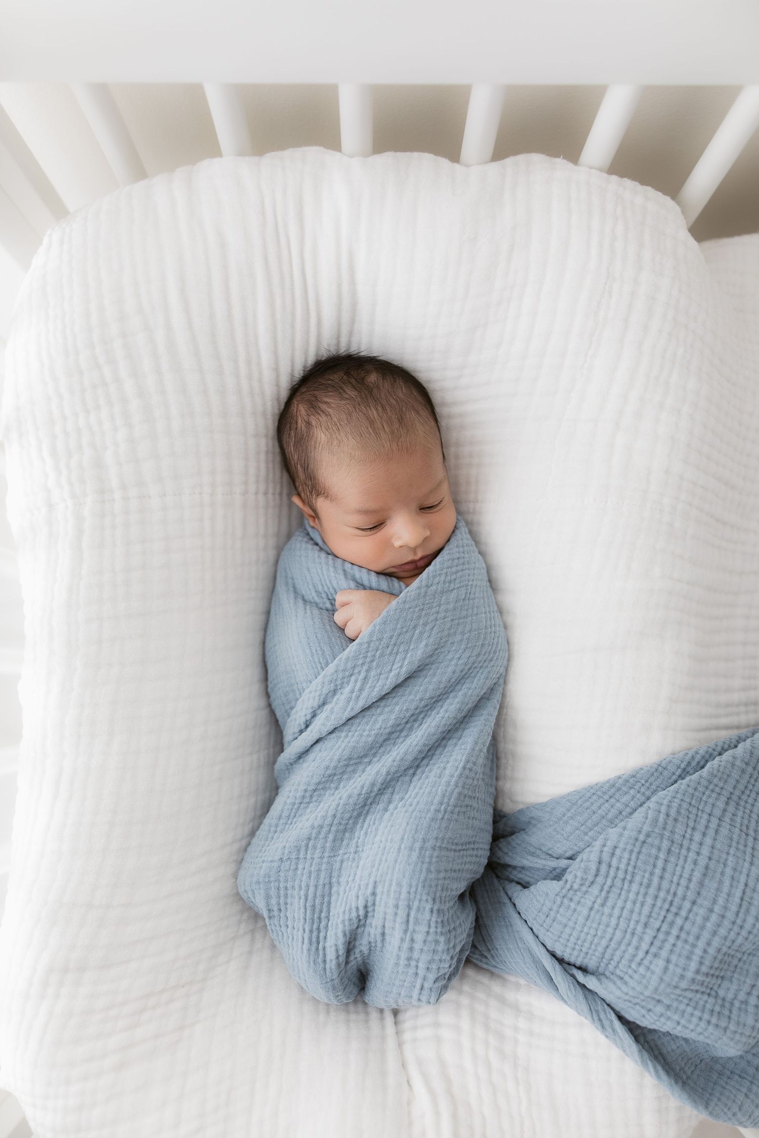 Sleeping newborn wrapped in blue swaddle on white lounger pillow.