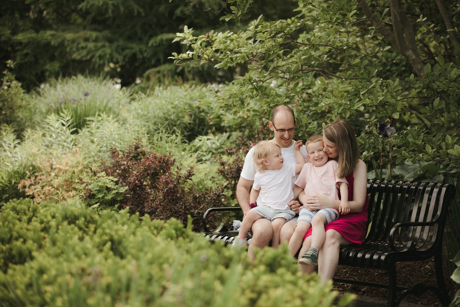 Family of four sitting on a bench surrounded by lush green garden shrubs, sharing a warm moment outdoors.