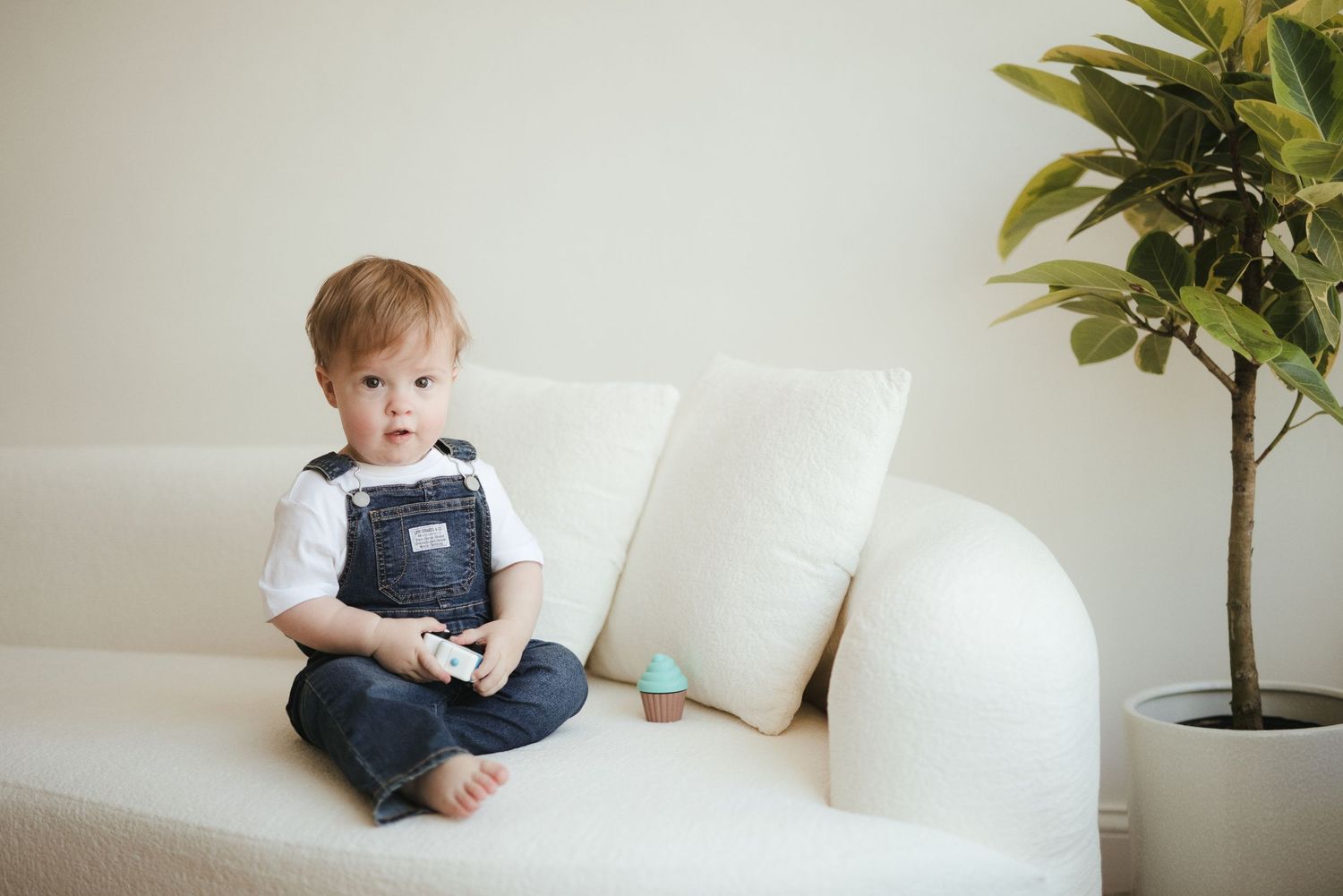 Toddler boy in denim overalls sitting cross-legged on a white sofa holding a toy, with a green plant nearby.