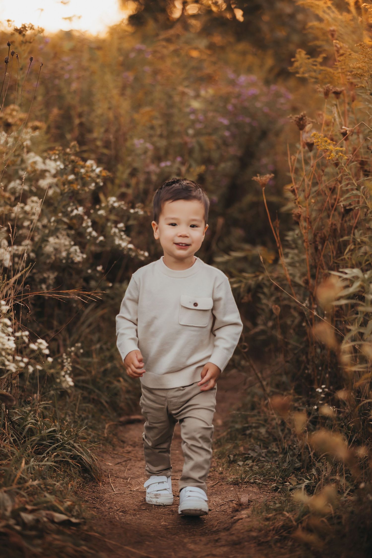 Toddler boy in beige outfit walking on a dirt path surrounded by wildflowers during golden hour.