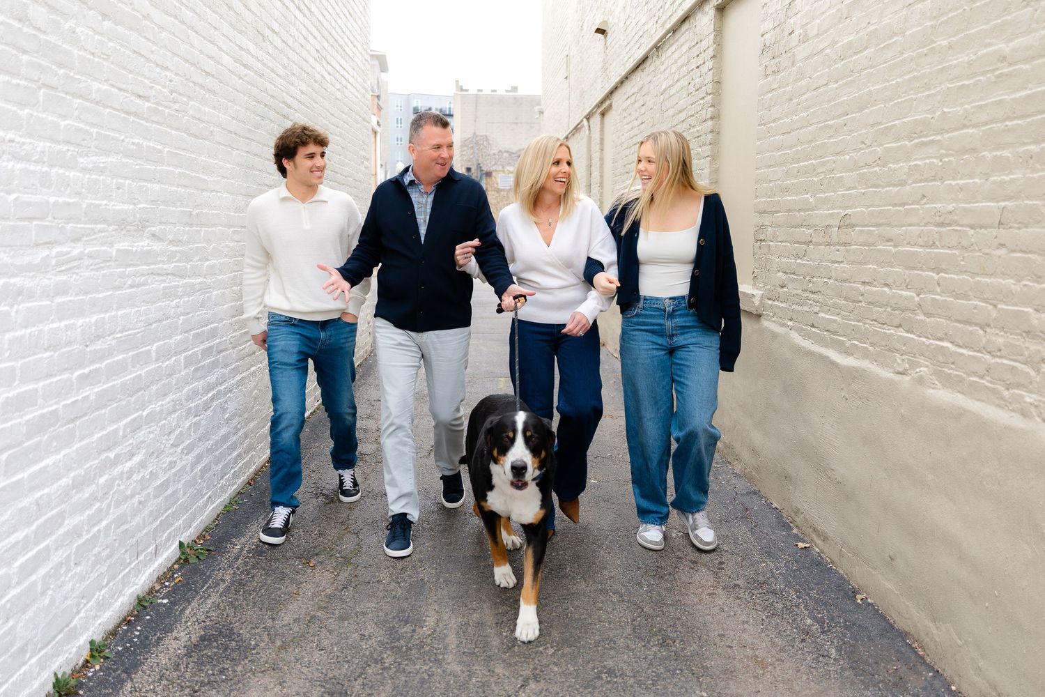 Family and their dog pose casually against white brick wall in matching casual outfits.