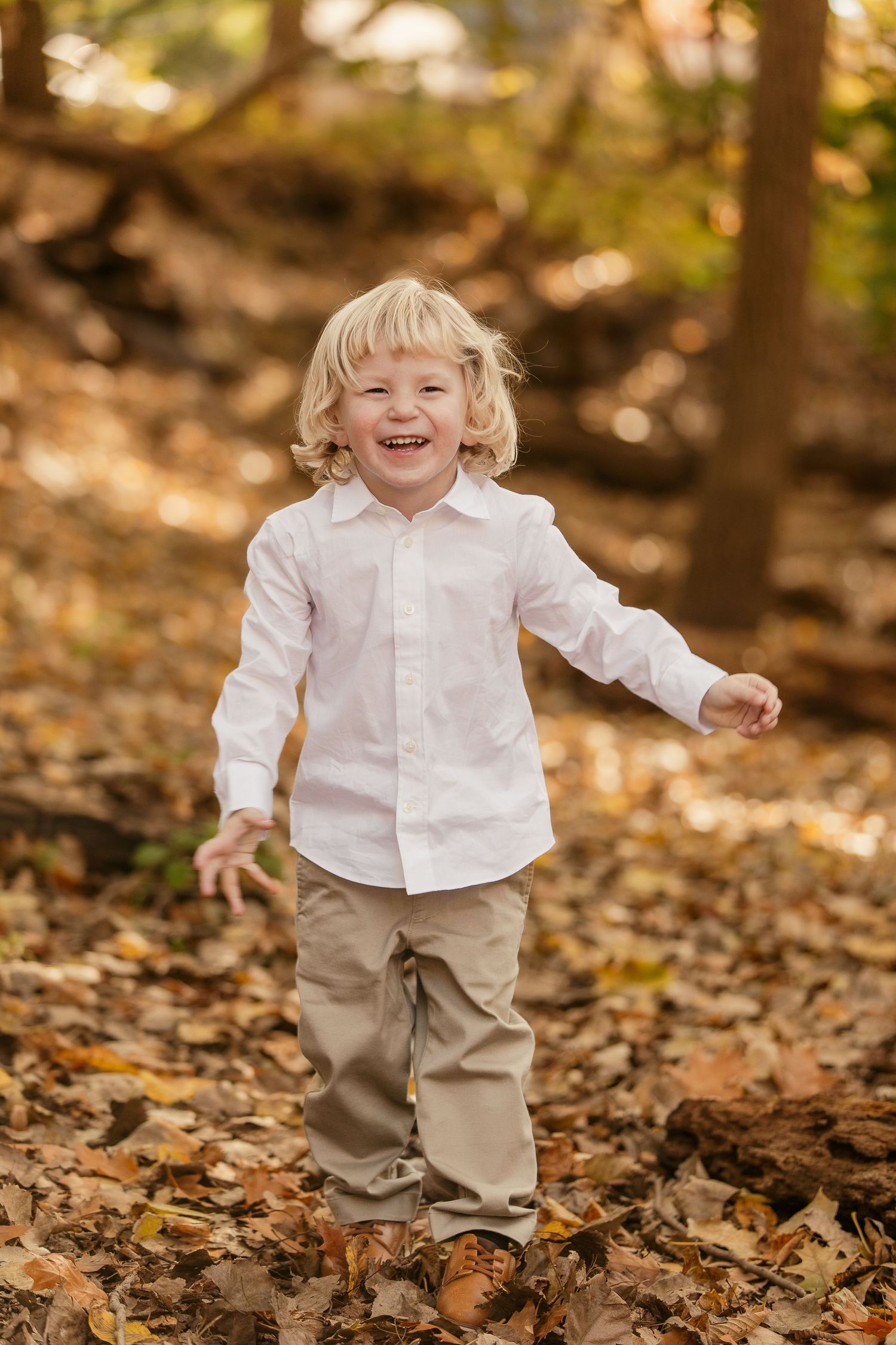 A joyful child in a white shirt and khaki pants plays in autumn leaves on a forest floor.
