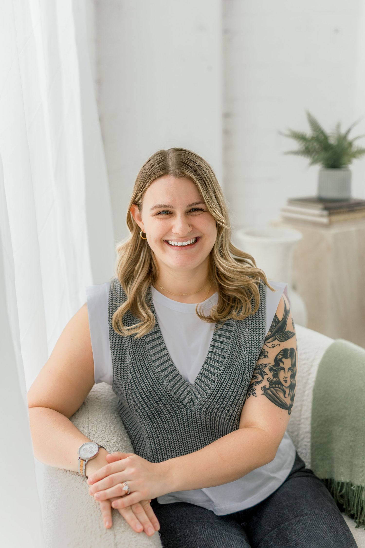 Professional headshot of person in gray sweater vest with visible arm tattoo sitting on white couch.