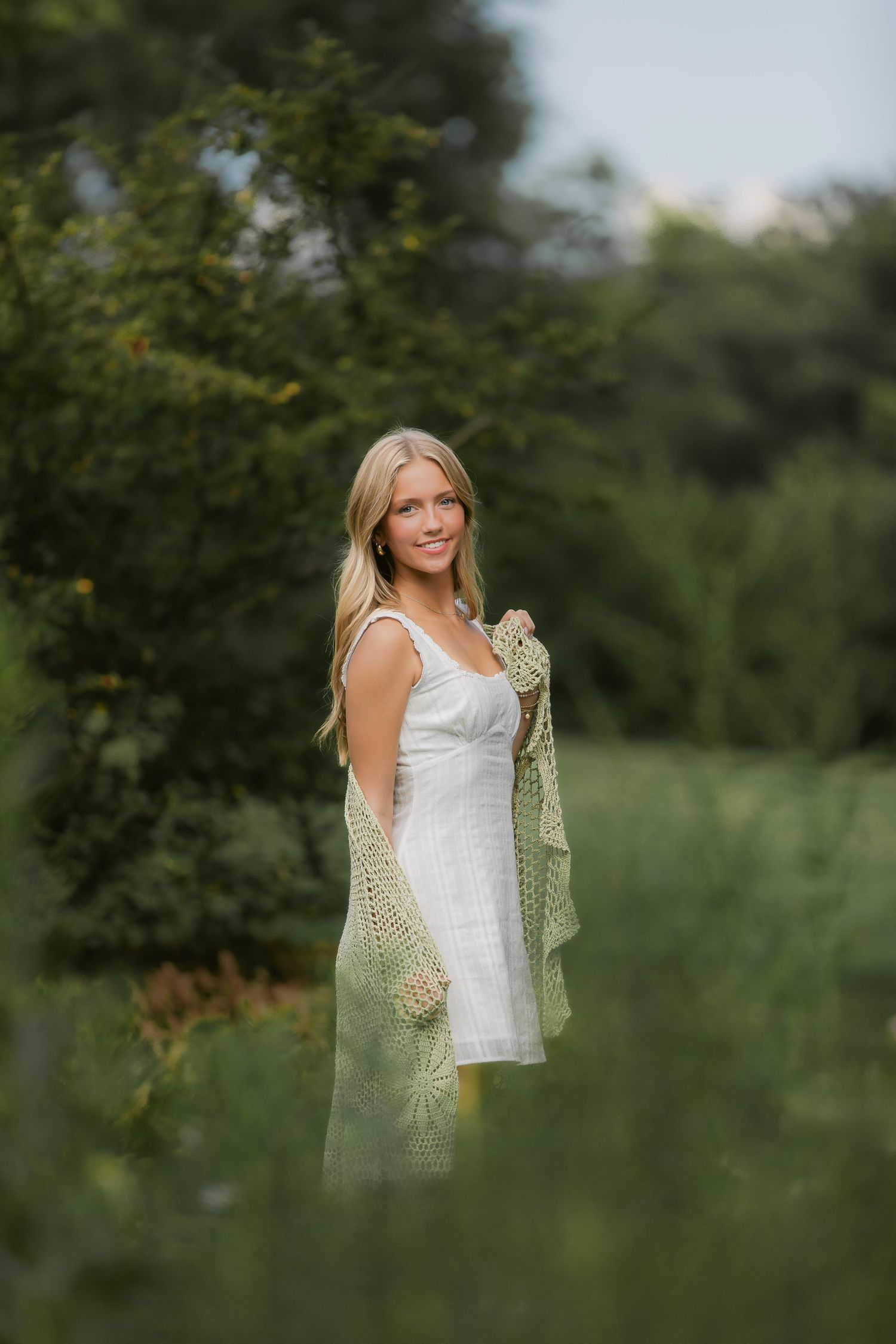 A figure in a white dress and green shawl stands elegantly in a lush summer garden.