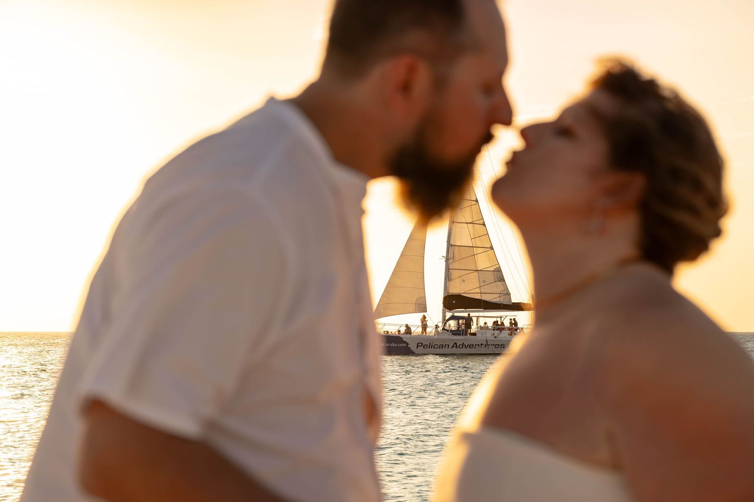 Couple sharing a romantic kiss at sunset with a sailboat on the ocean in the background.
