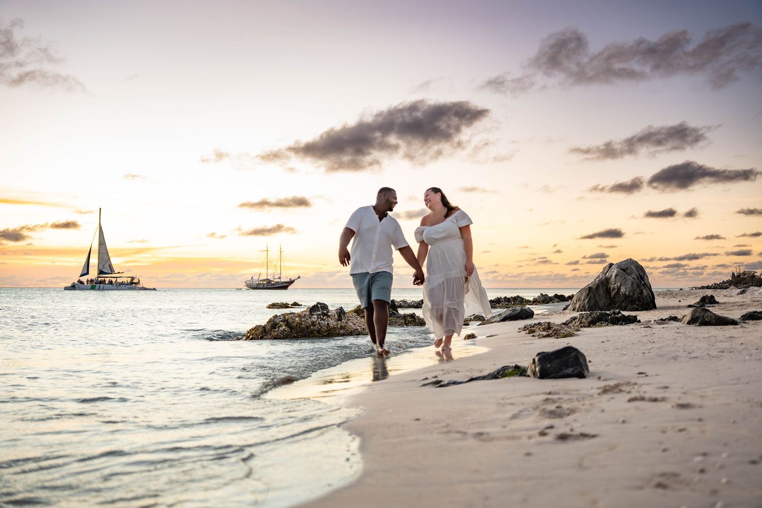 Couple holding hands walking on a tropical beach at sunset with sailboats in the background.
