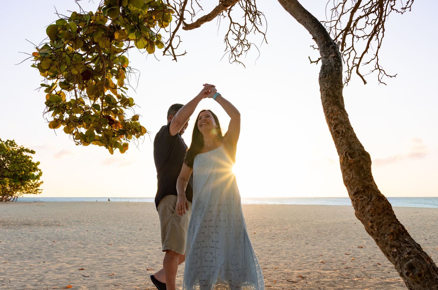 A couple dances together on a beach at sunset under a leaning tree with golden sunlight streaming through the branches.