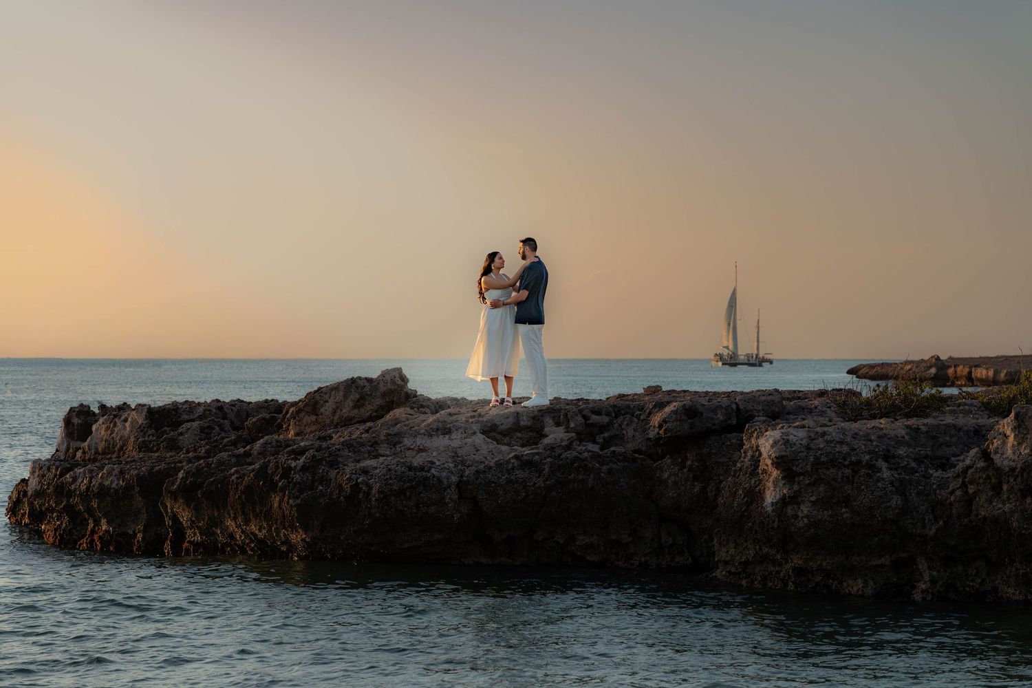 Couple embracing on rocky shoreline at sunset with a sailboat on the horizon during a romantic engagement photo session.