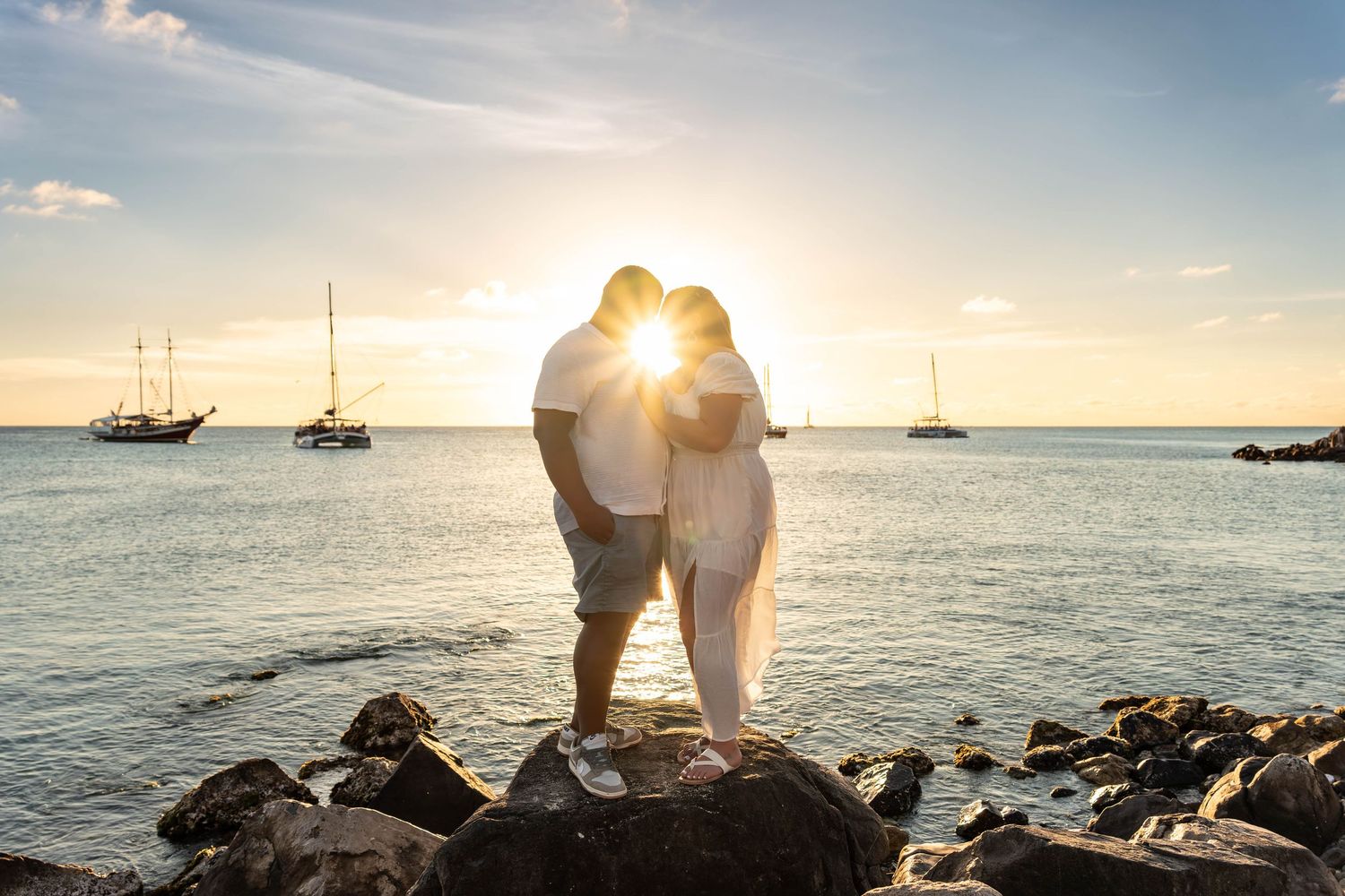 Couple kissing on rocks at sunset by the ocean with sailboats in the background.