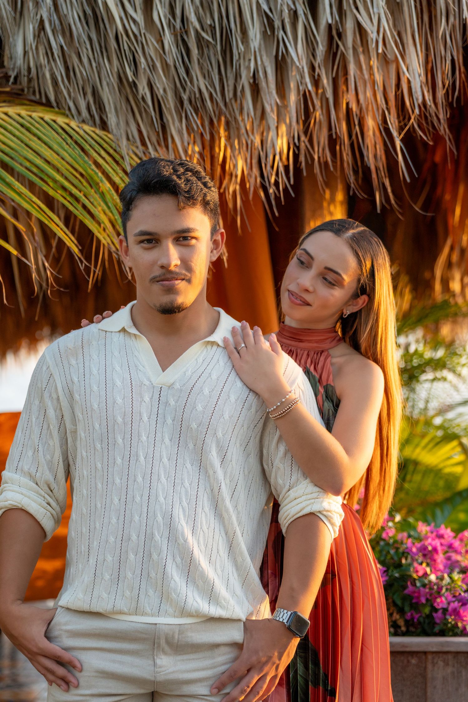 A couple poses together under a thatched palapa roof with tropical flowers and palm trees in the background.