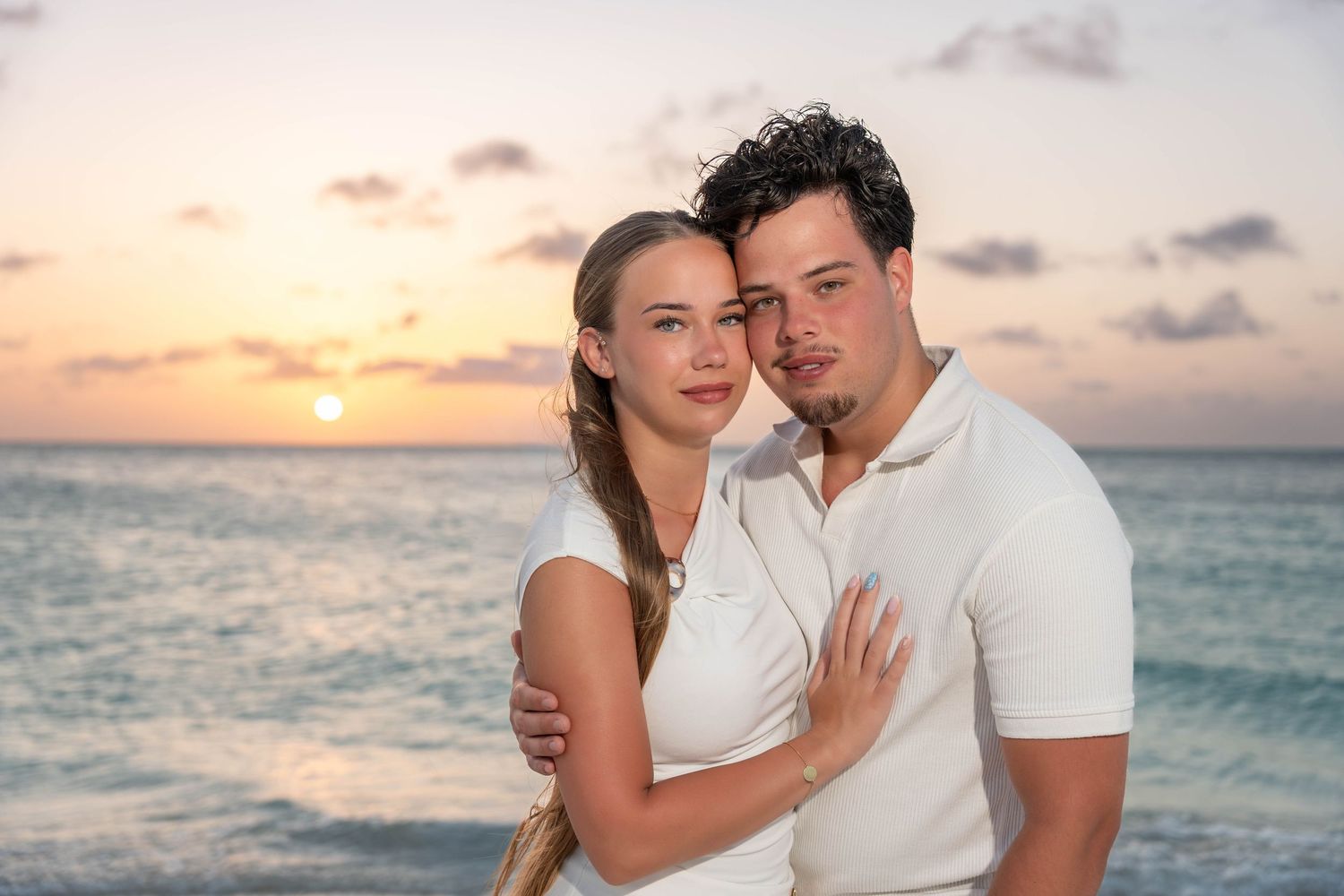 Couple embracing at sunset on a tropical beach, both wearing white outfits with ocean waves in the background.