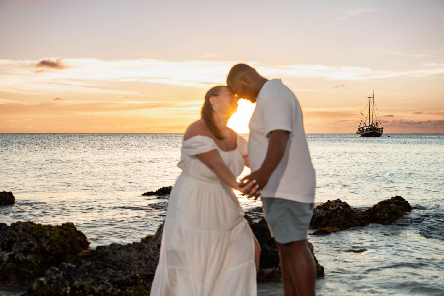 Couple sharing a romantic sunset moment by the ocean, with a sailing ship in the background during golden hour.