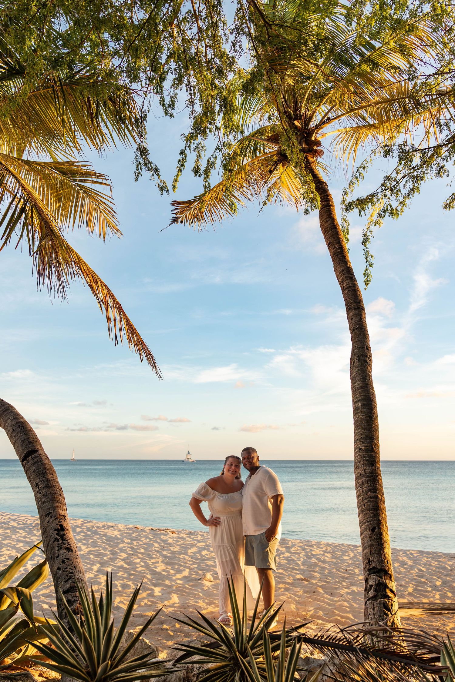 Couple embracing on a tropical beach at sunset, framed by palm trees with calm turquoise ocean in the background.