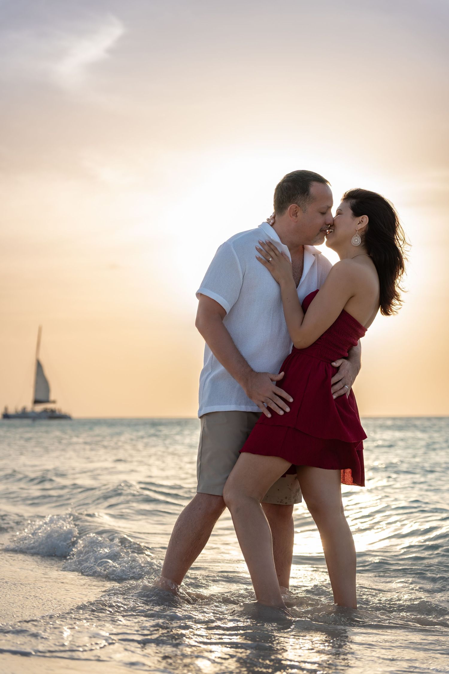 Romantic couple kissing at sunset on a tropical beach, standing in shallow waves with a sailboat in the background.