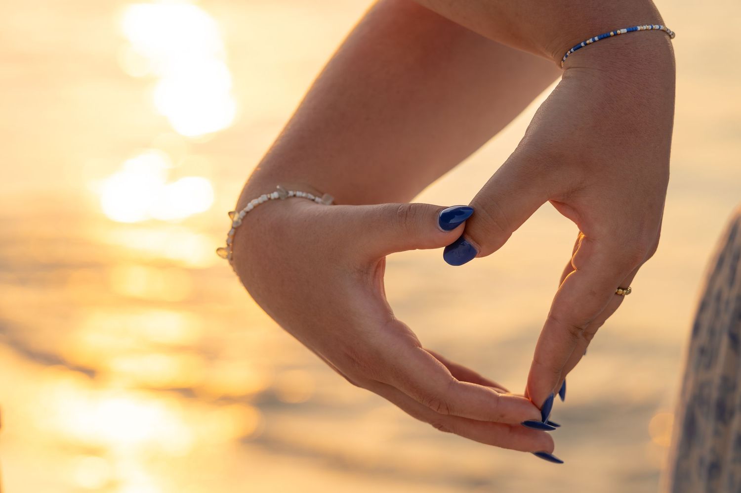 Hand with blue nail polish reaches toward golden sunset over ocean horizon creating silhouette against warm sky.