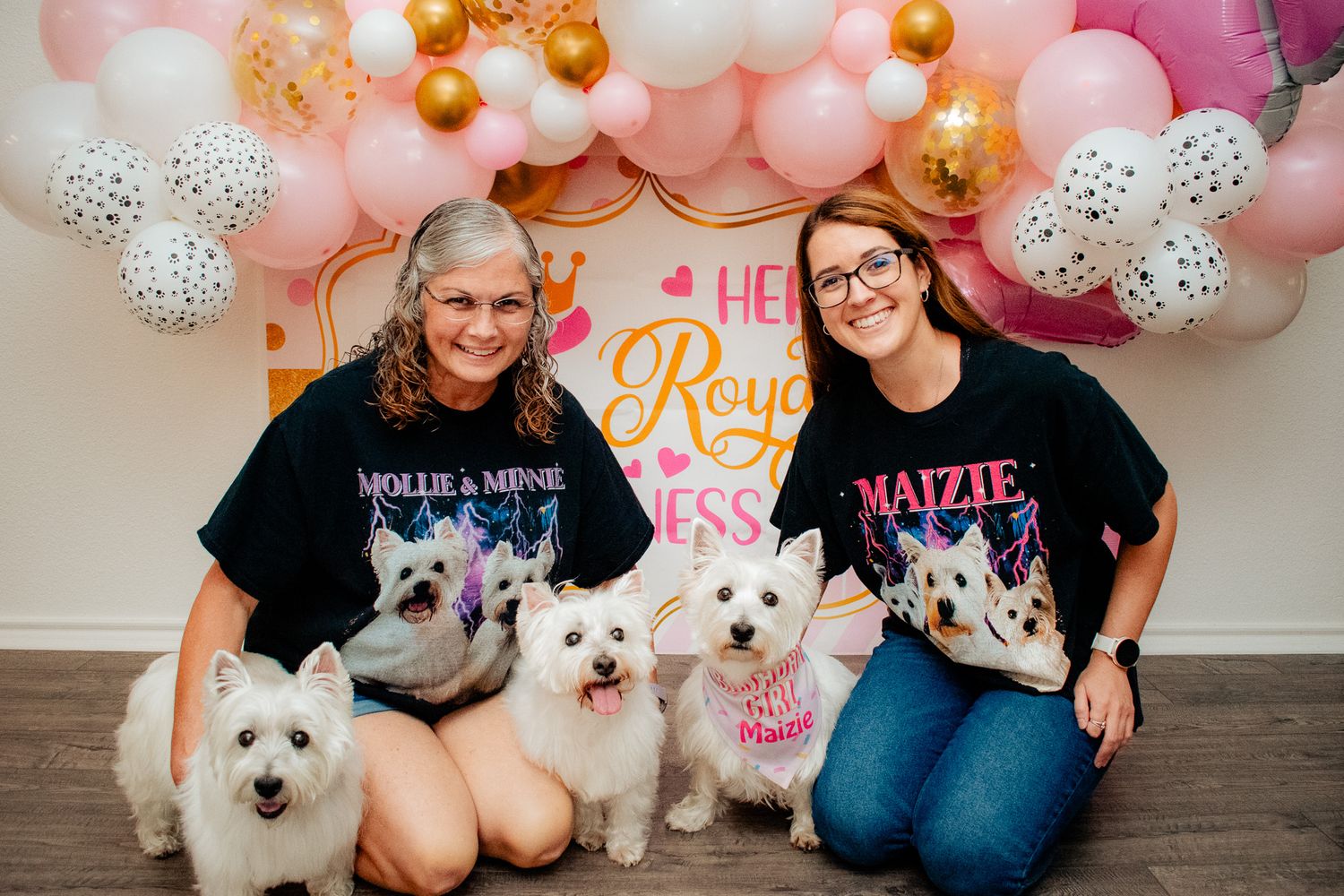 White dogs pose with their owners at a pink and gold balloon-decorated celebration.