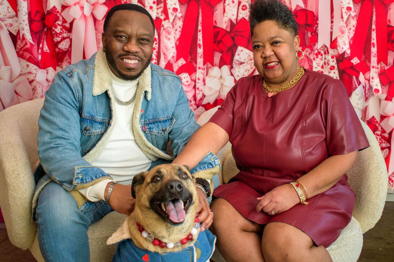 Two people sitting on chairs with their pug dog against a red and white patterned background.