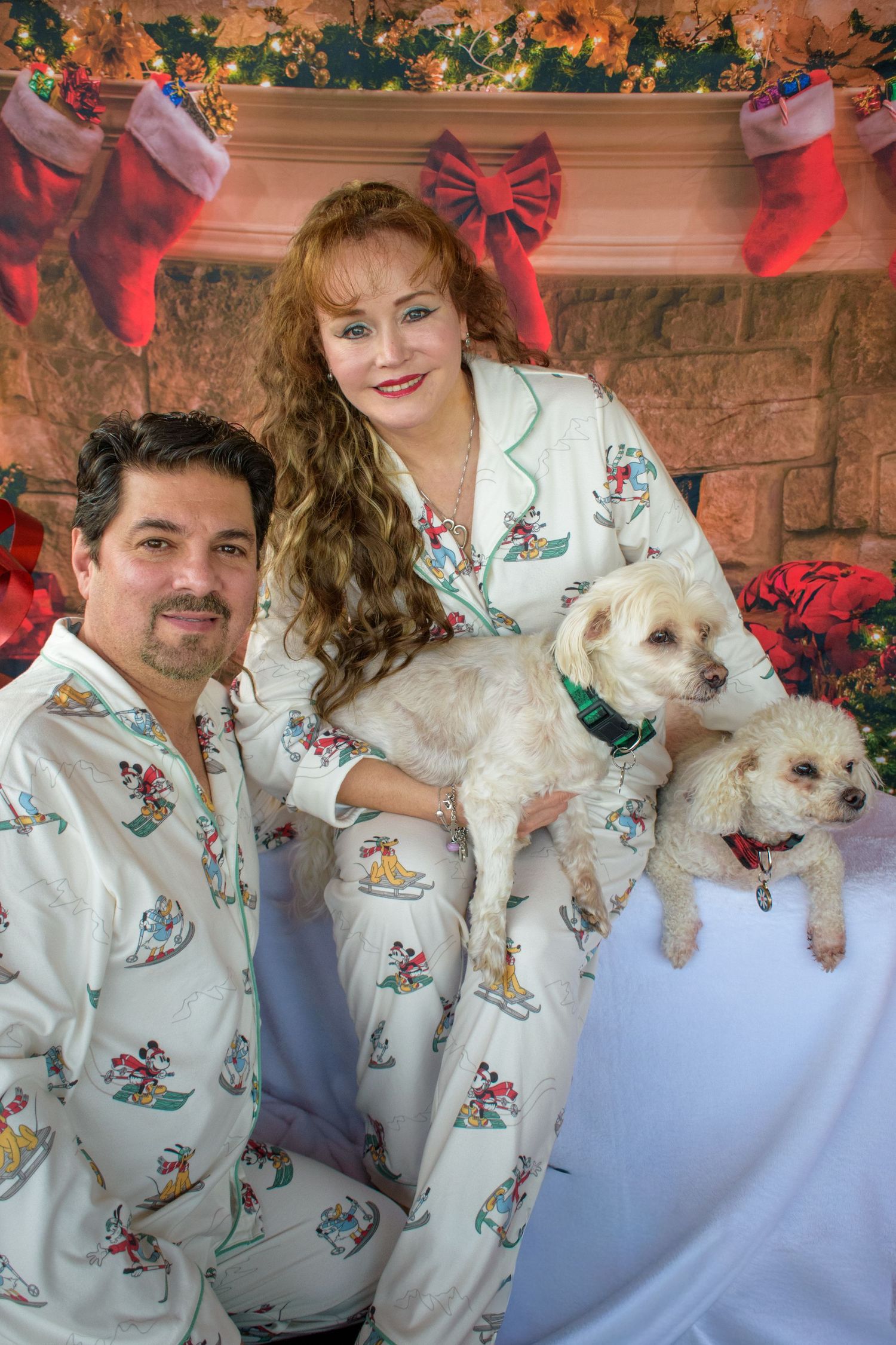 Couple in matching white holiday sweaters posing with small dogs against festive backdrop.