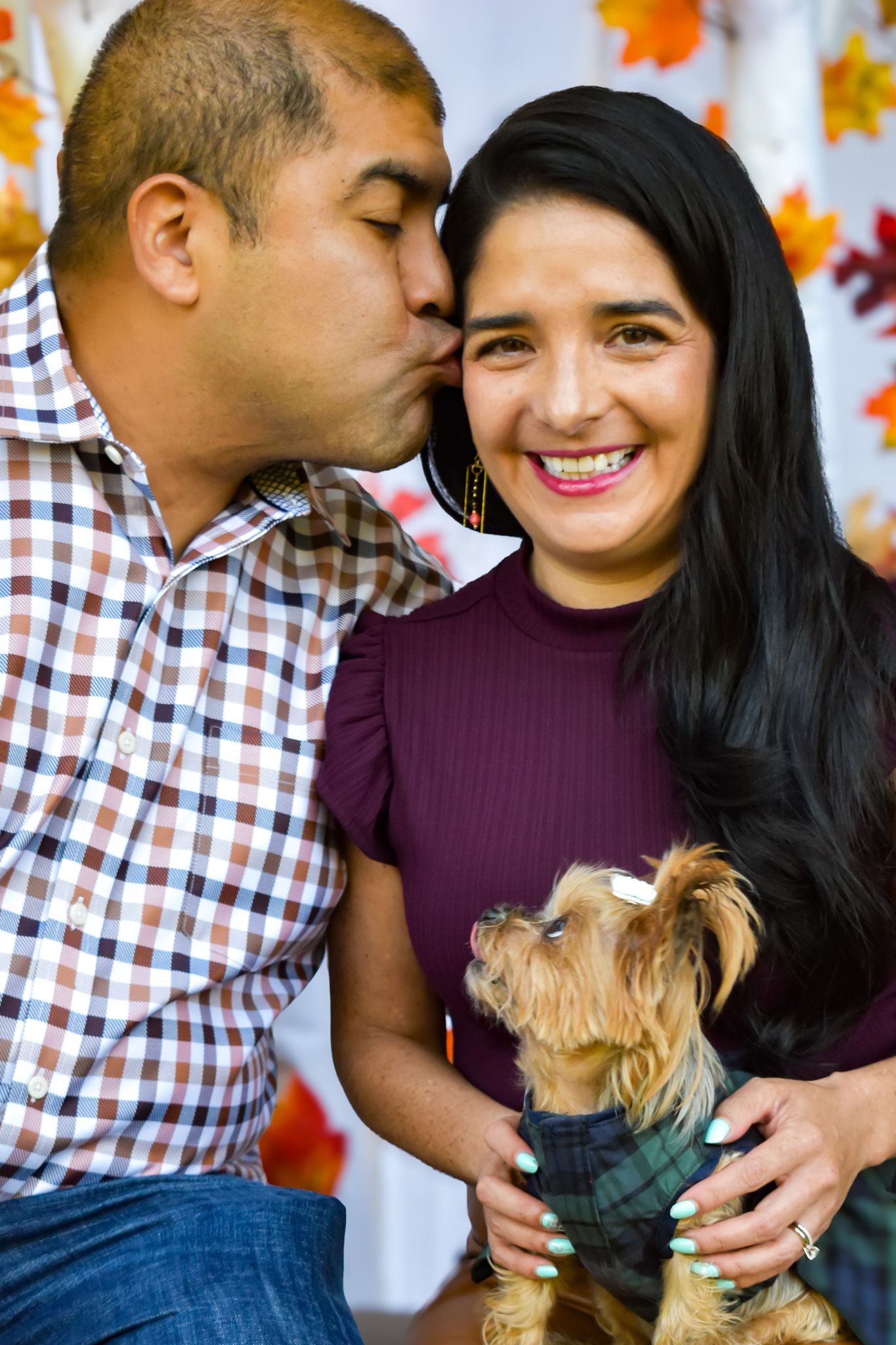 A couple shares intimate moments with their small dog against a fall-themed backdrop.