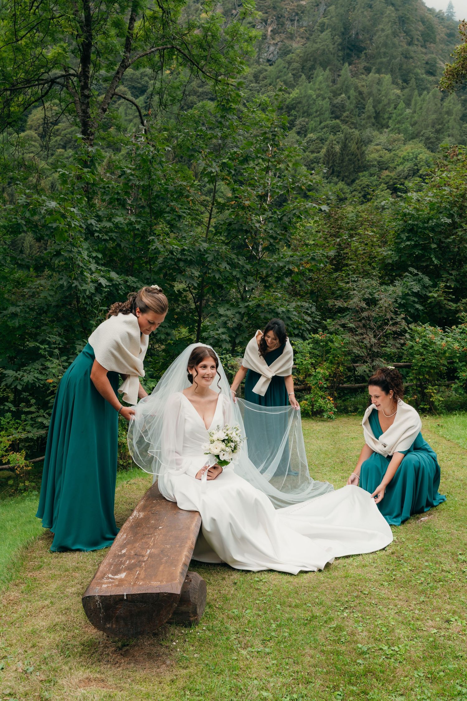 Bride in white gown sits on wooden bench while three bridesmaids in teal dresses arrange her veil outdoors.
