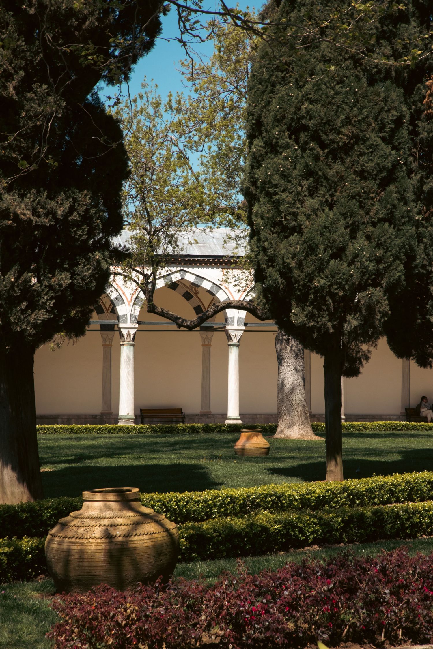 Lush garden courtyard with cypress trees, decorative urns, and arched colonnades of a historic Ottoman building.