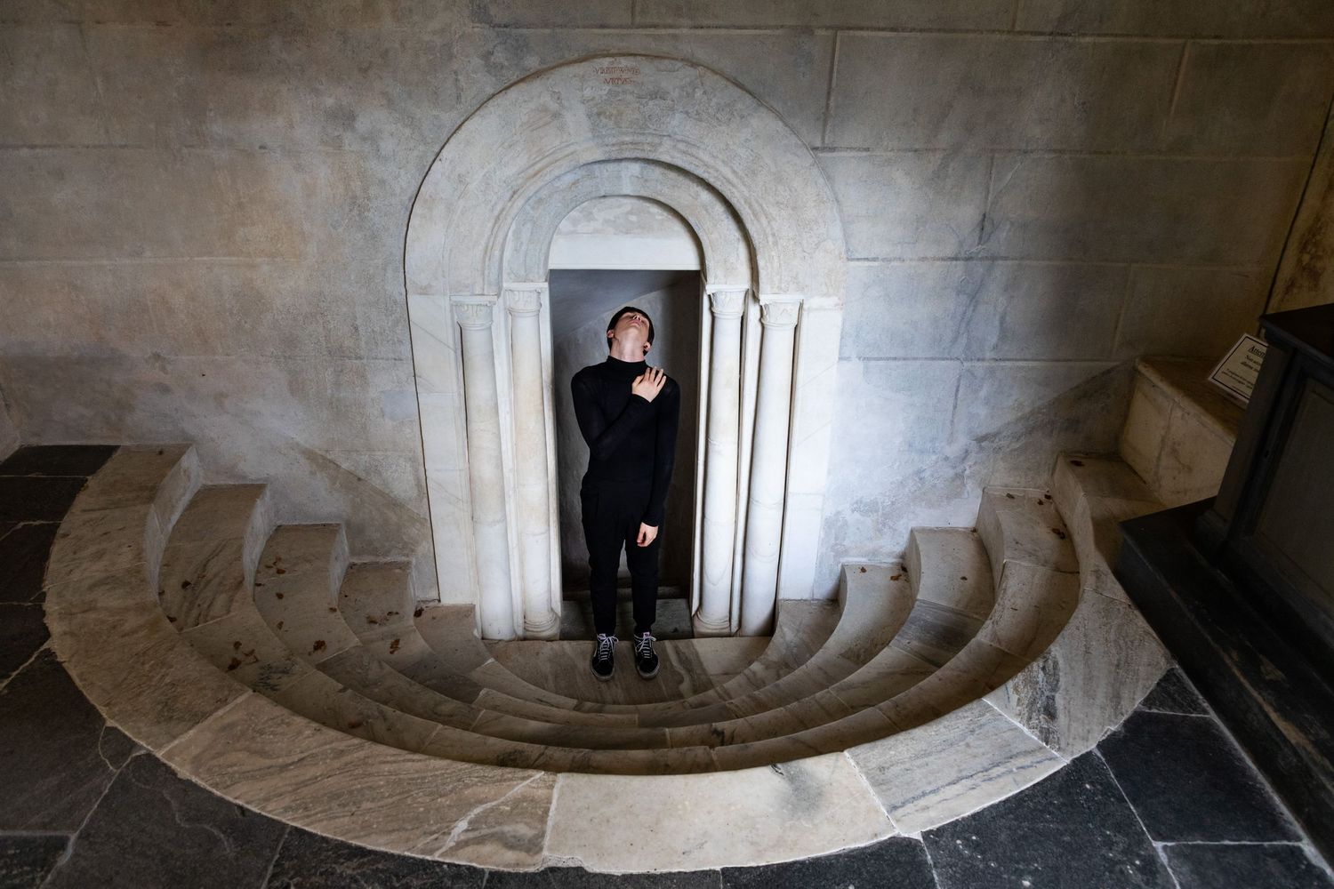 Person standing in arched doorway at bottom of curved marble staircase in elegant stone building.