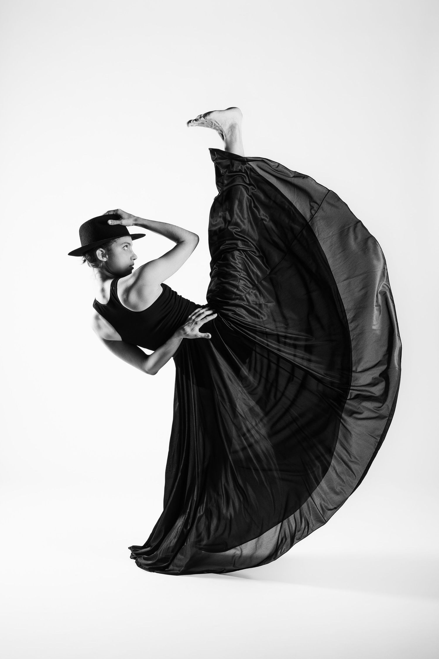 A dancer in a black cap and flowing black skirt performs a dramatic backbend in a black and white studio photo.