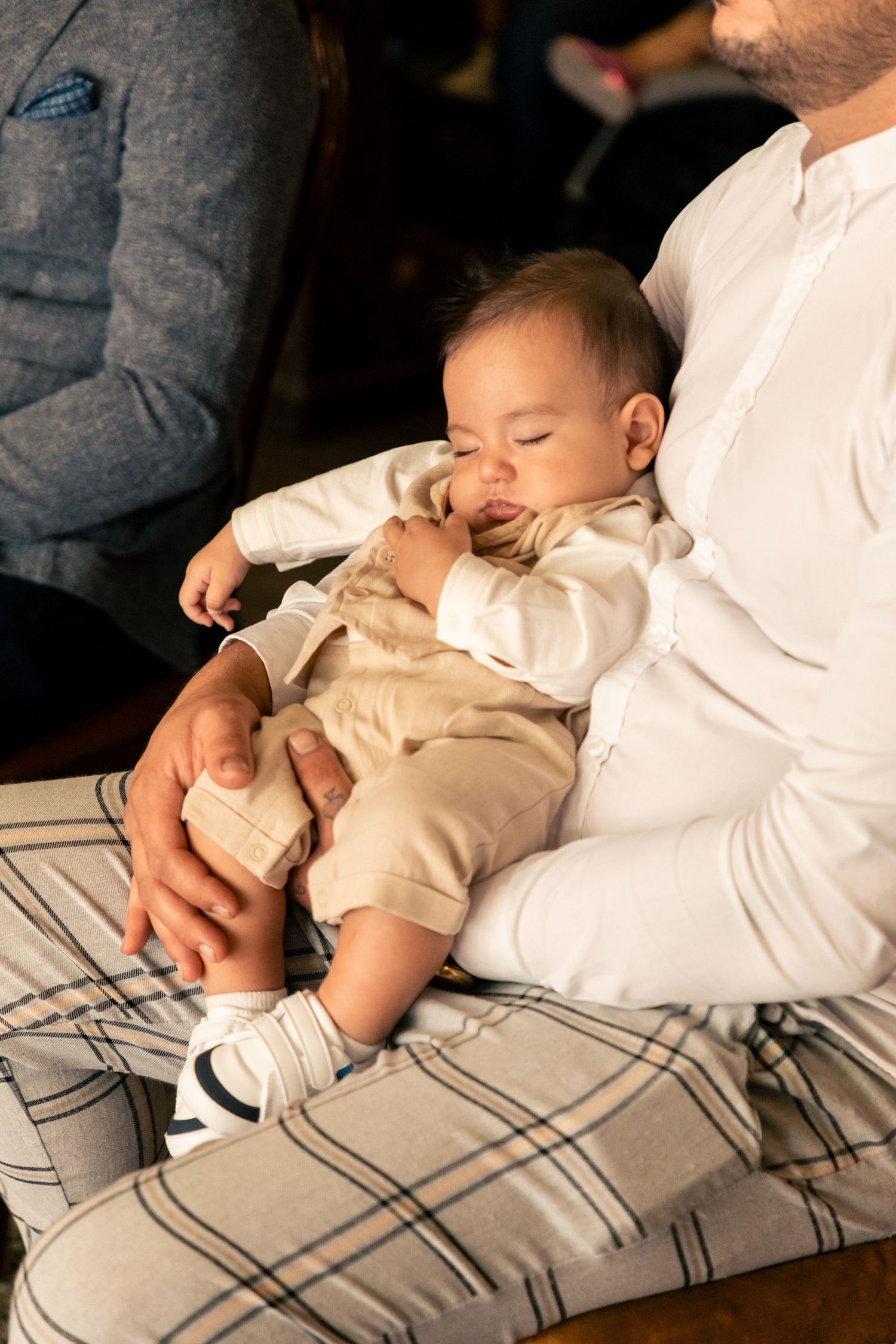 Sleeping newborn baby in beige outfit resting on fathers lap during baptism ceremony.