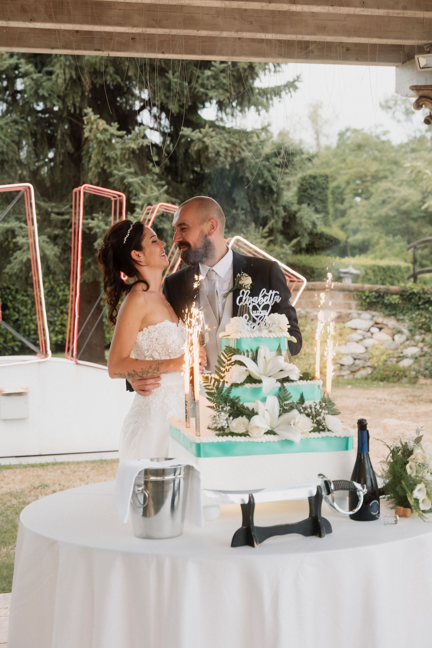Bride and groom share a kiss beside a teal tiered wedding cake with sparklers outdoors.