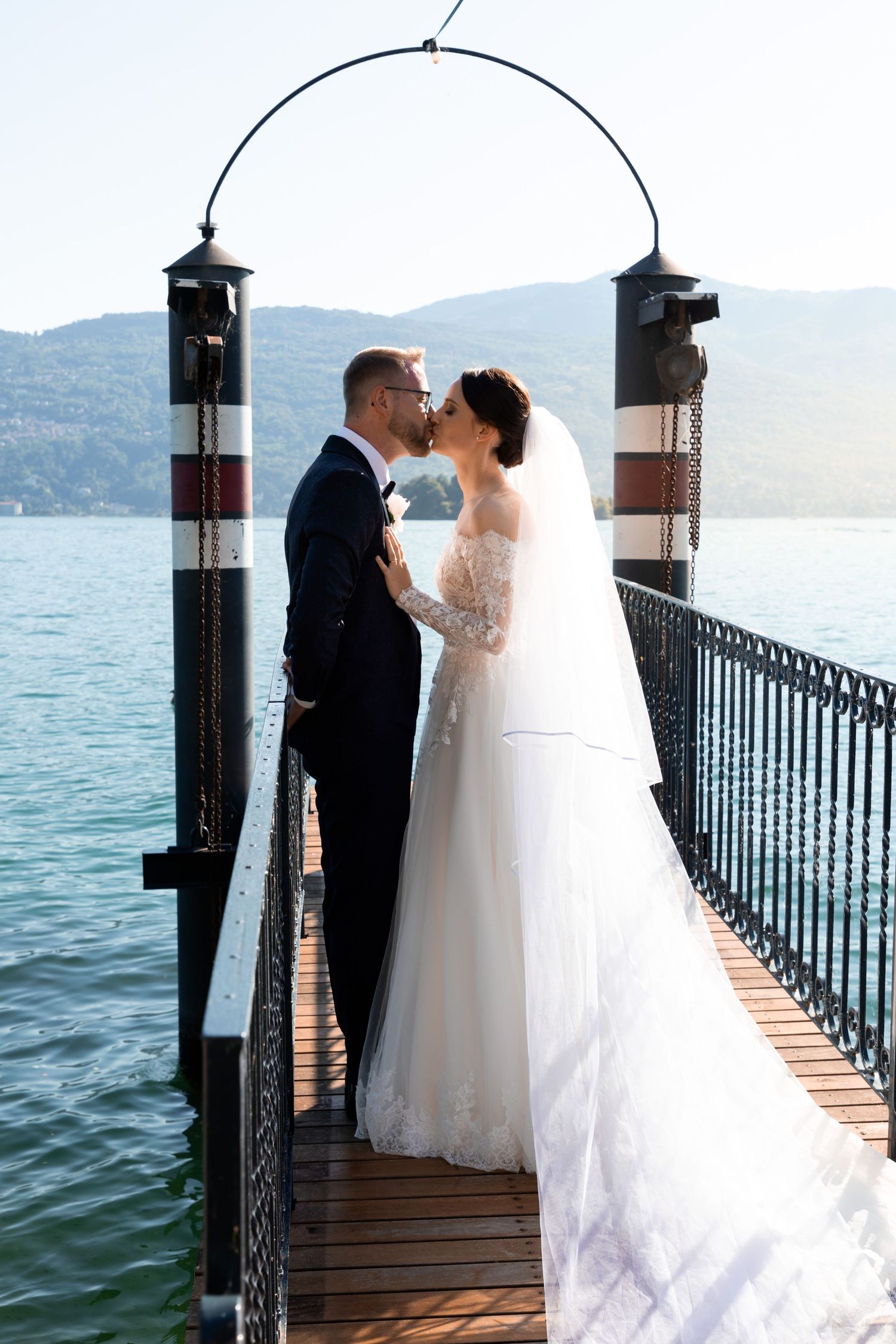 Wedding couple shares romantic kiss on wooden pier with blue lake and mountain backdrop on sunny day.