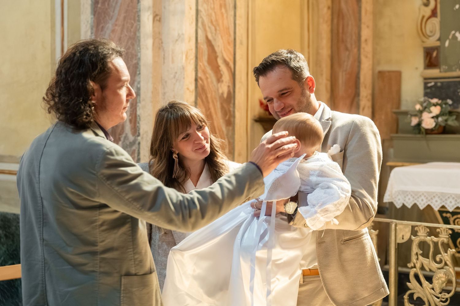 Godparents and father tenderly hold a baby in white christening gown inside an ornate church.