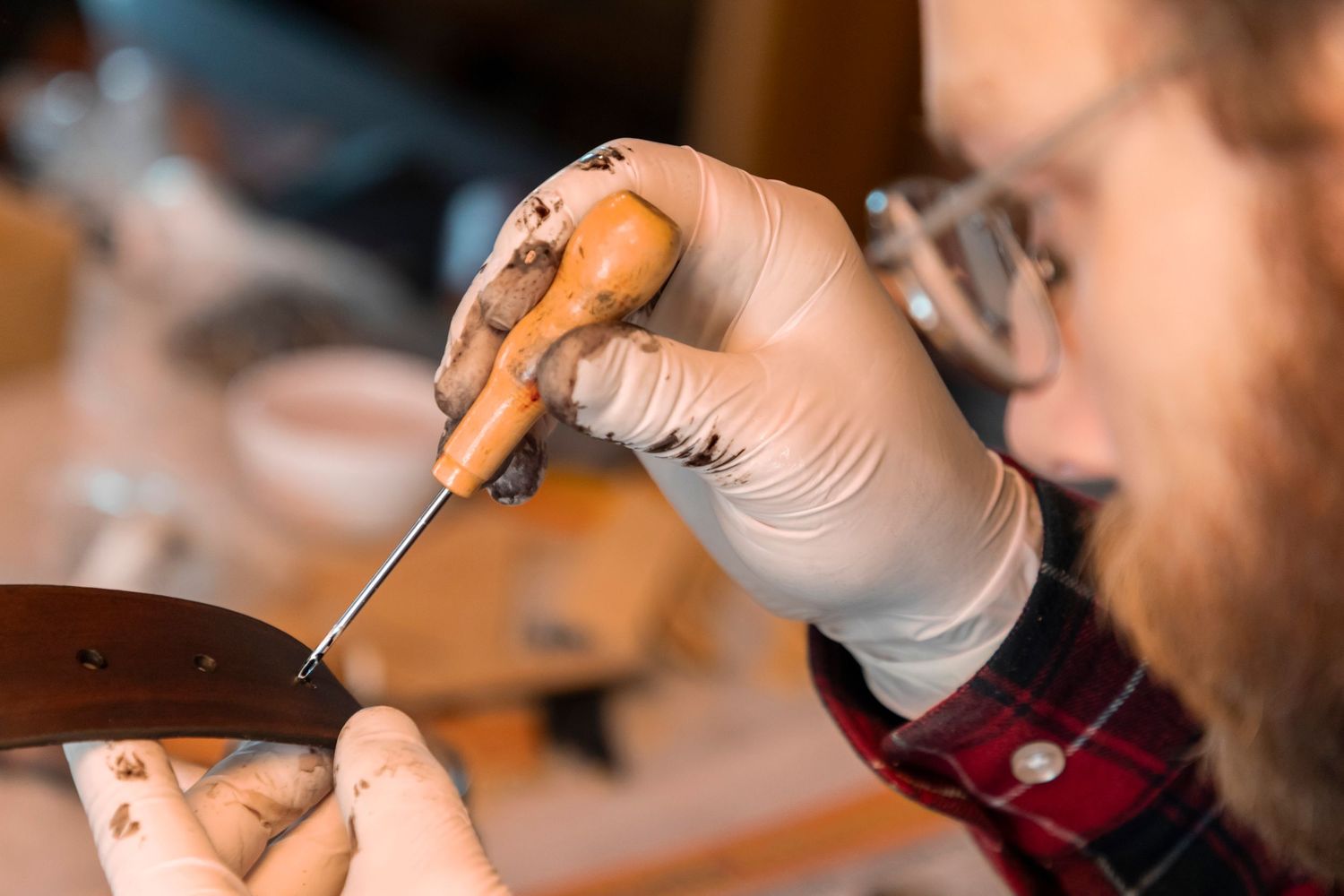 Bearded craftsman in gloves using awl tool to punch holes in leather belt in workshop.