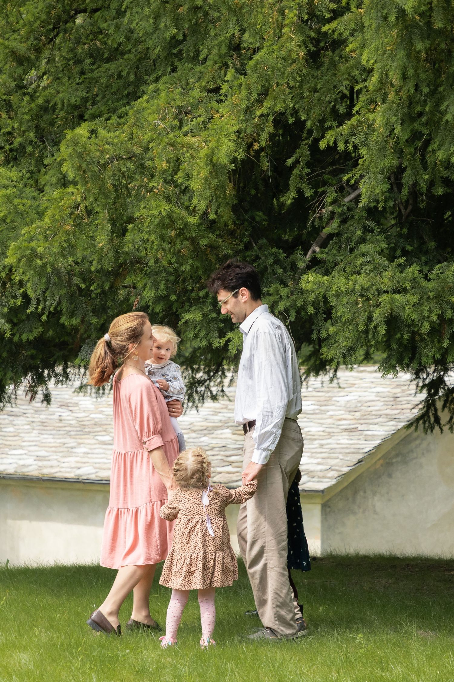 Young family with two children standing on green lawn near large tree.