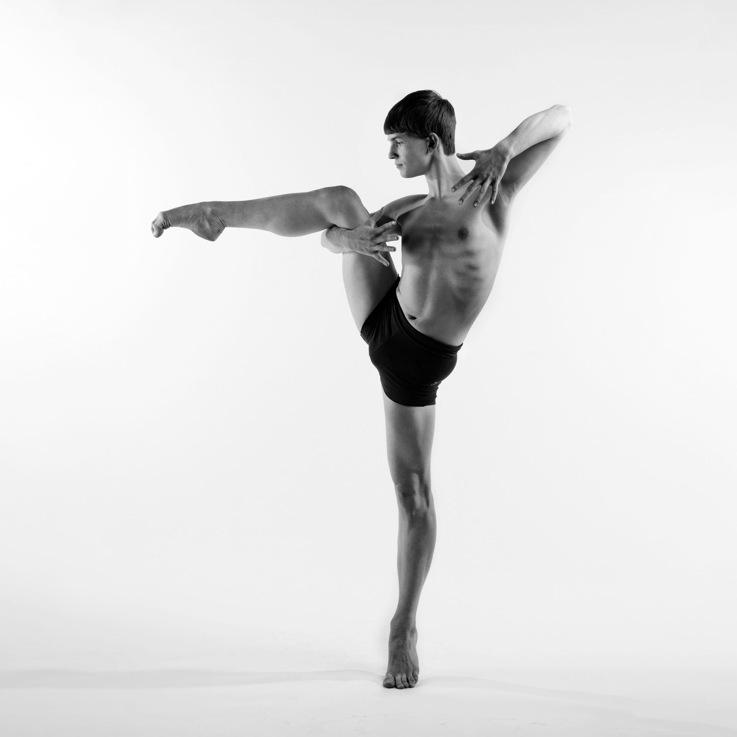 Athletic male ballet dancer balancing on one leg with high kick and extended arms in black shorts.
