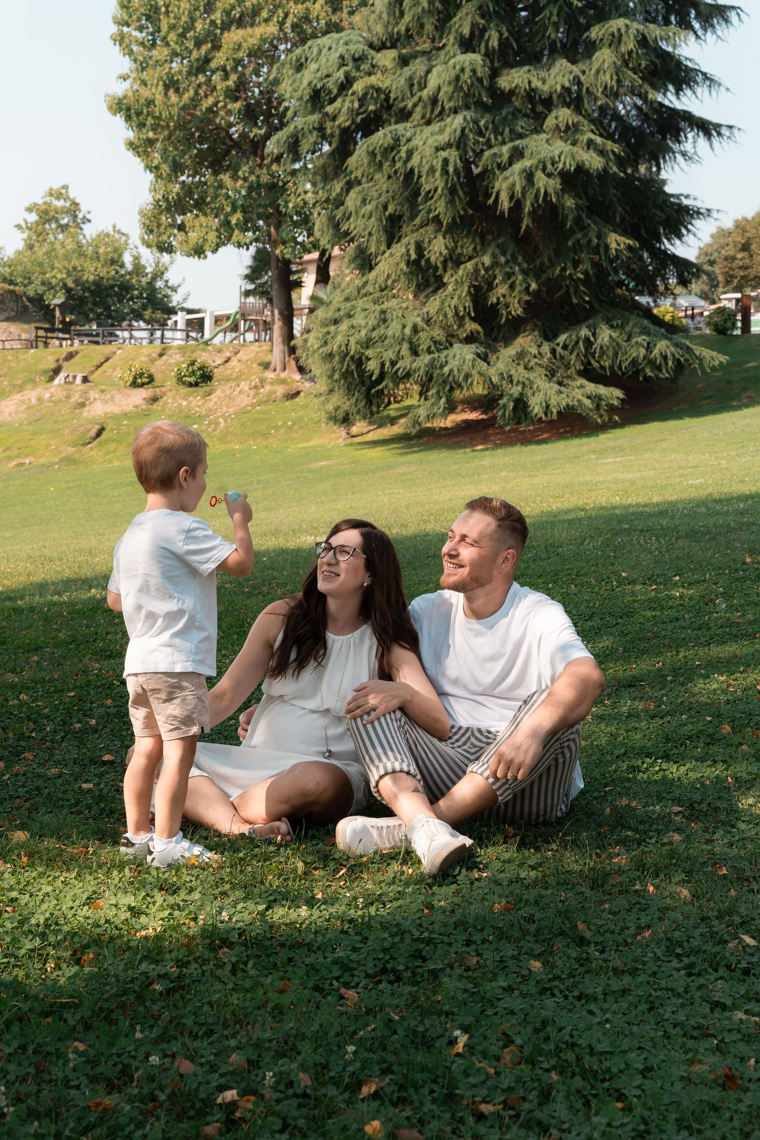 A young family sits on green grass in a sunny park while their toddler son shows them something.