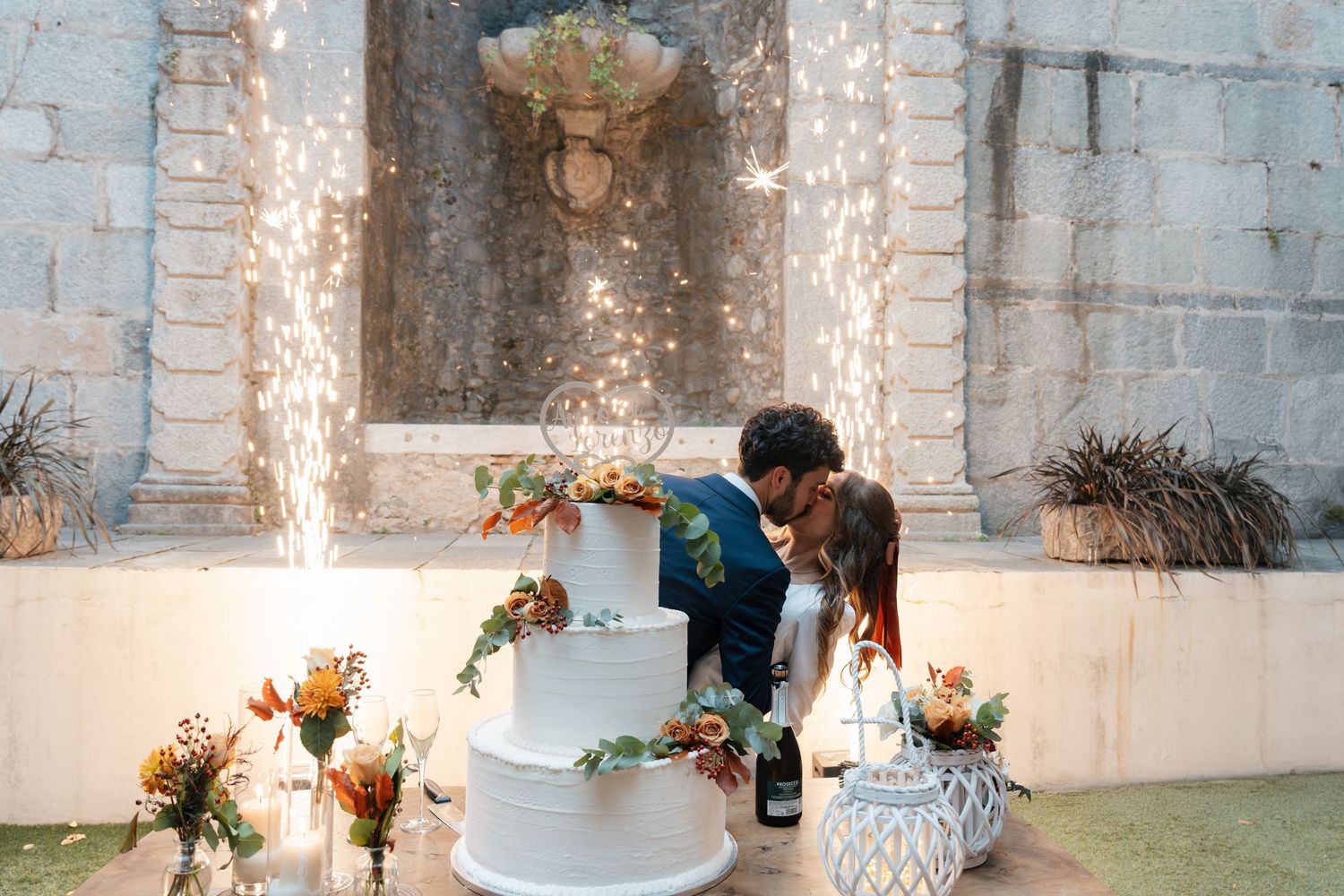 Bride and groom kiss behind white tiered wedding cake with sparkling fireworks display against stone wall backdrop.