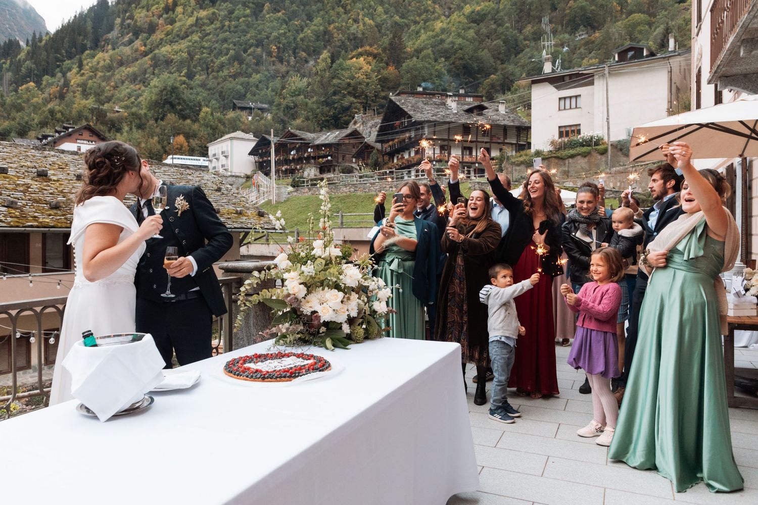 Wedding guests raising glasses in toast on outdoor mountain village terrace celebration.
