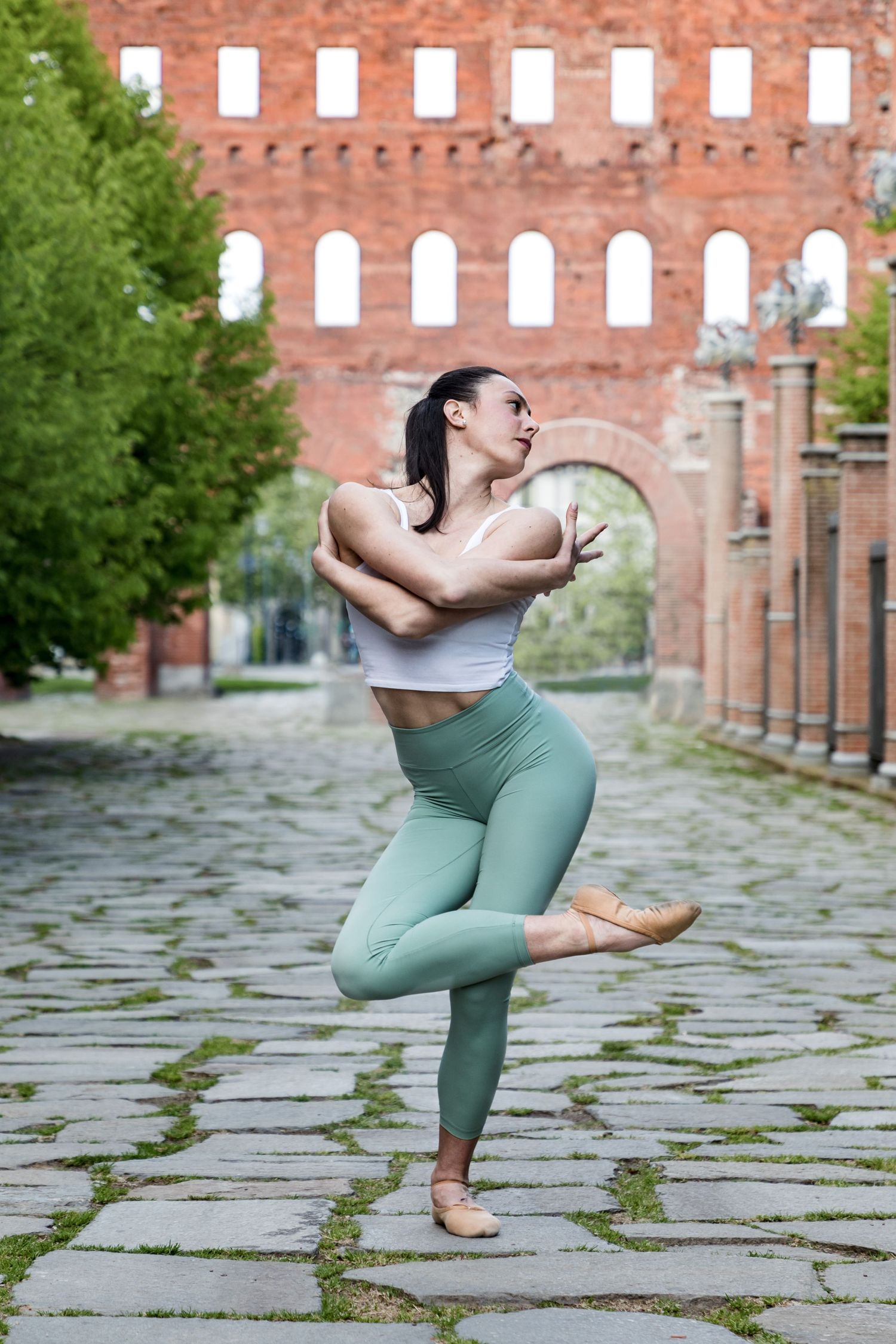 Female dancer in mint leggings performing balance pose on cobblestone street near brick arch.