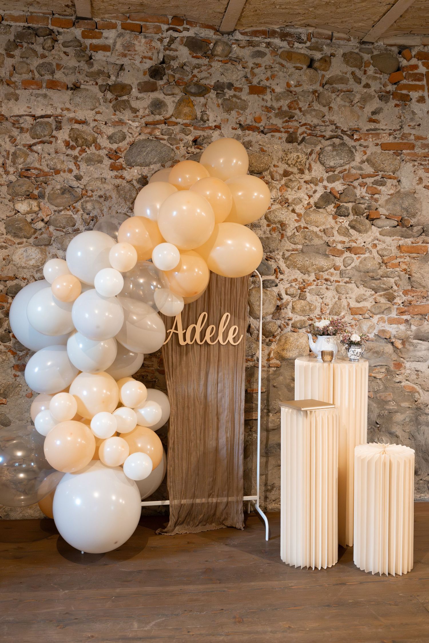 Elegant balloon arch with name Adele and wooden pedestals against rustic brick wall.