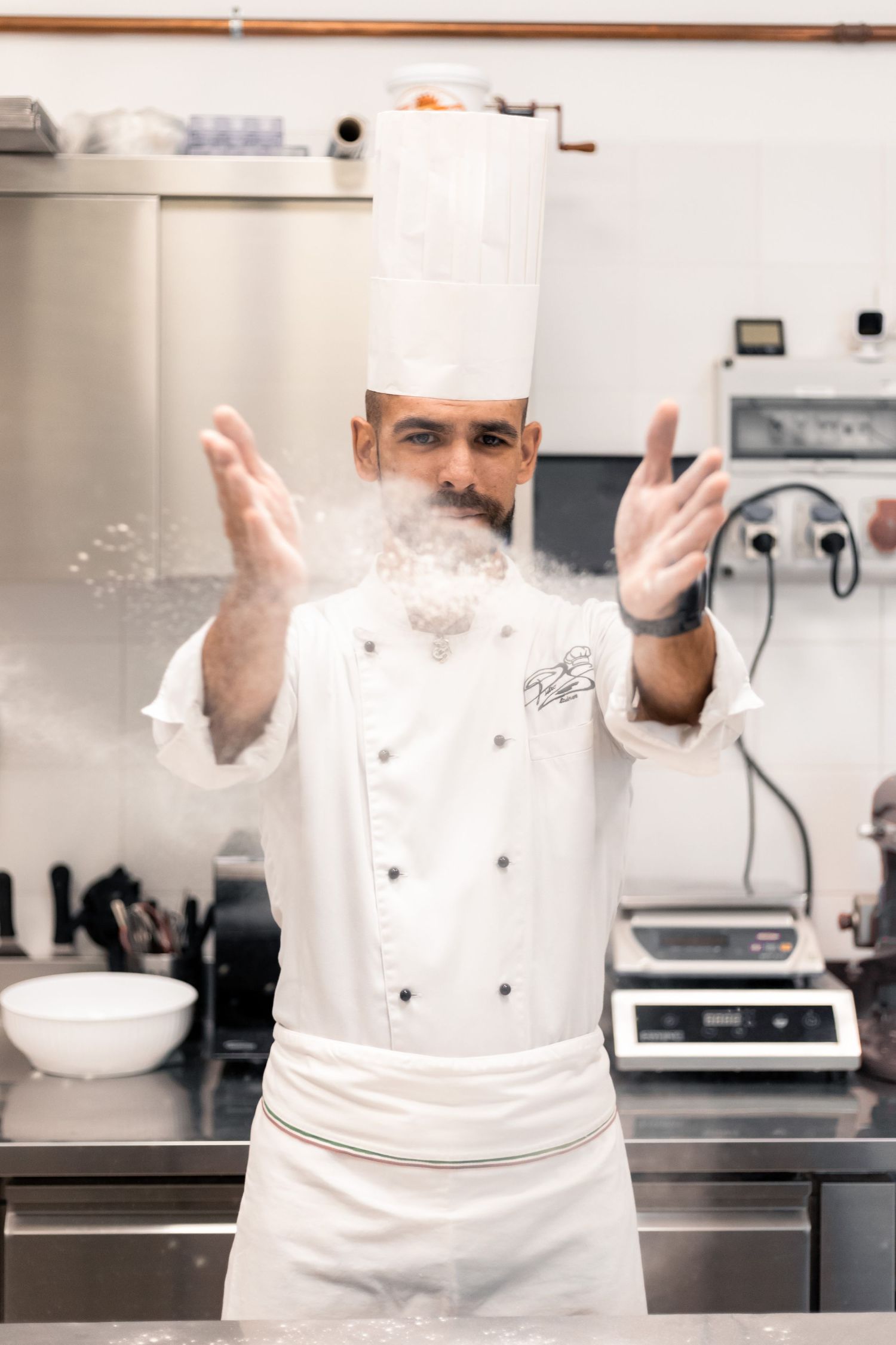 A bearded chef in white uniform clapping flour from his hands in a kitchen.
