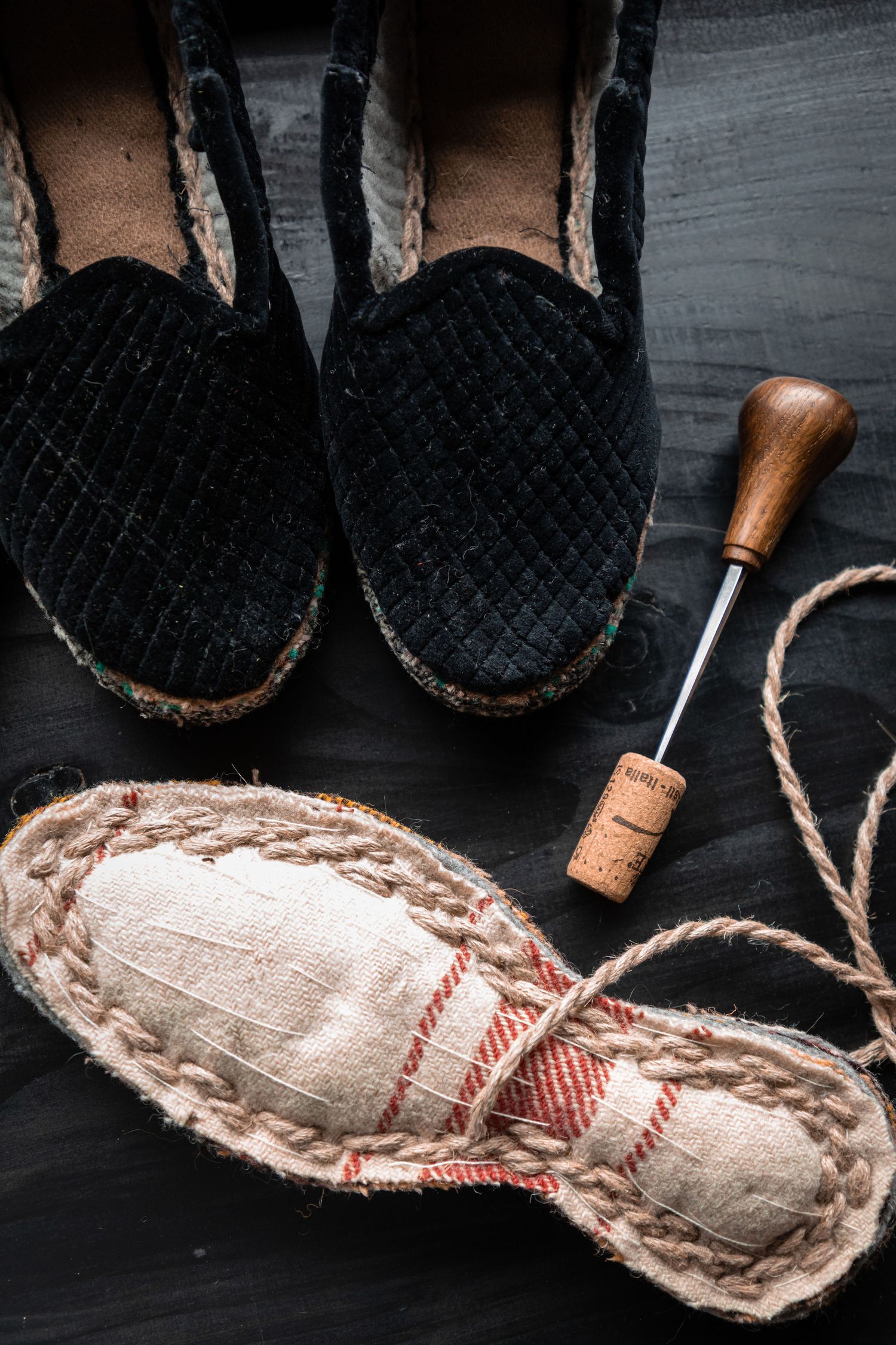 Overhead view of black velvet espadrilles beside an unfinished sole, needle, and cork tool on dark wood.