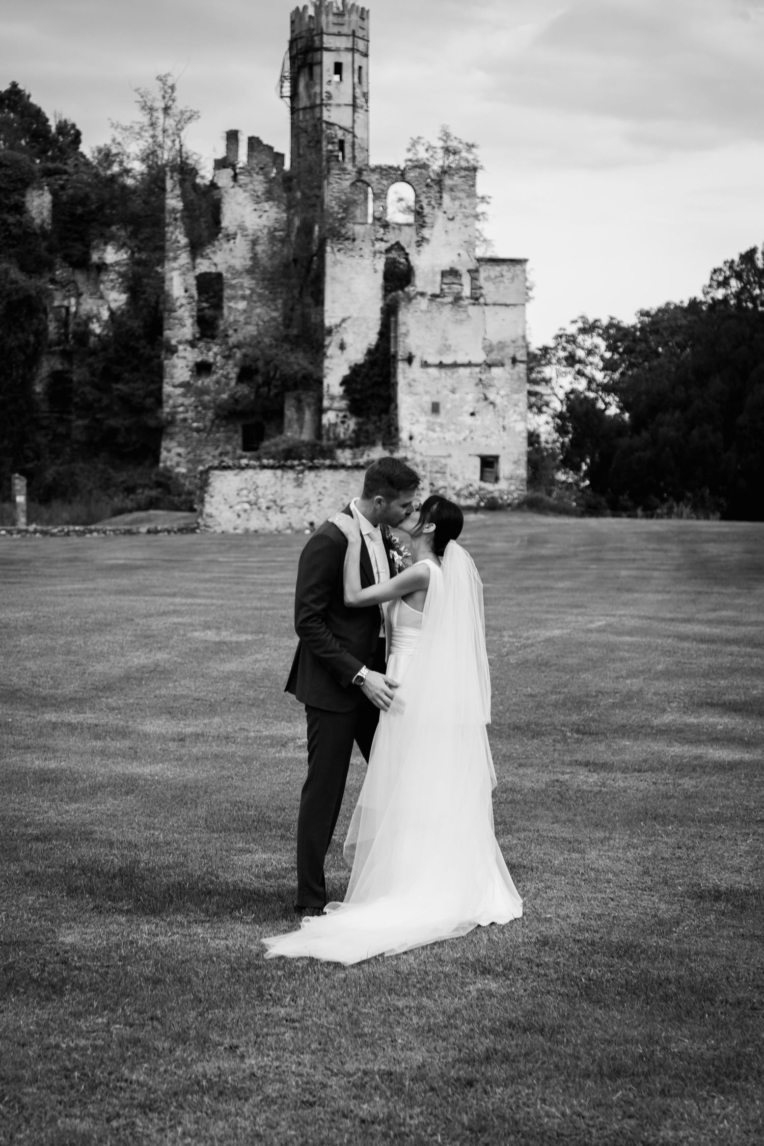 Black and white photo of bride and groom kissing on lawn before historic castle ruins.