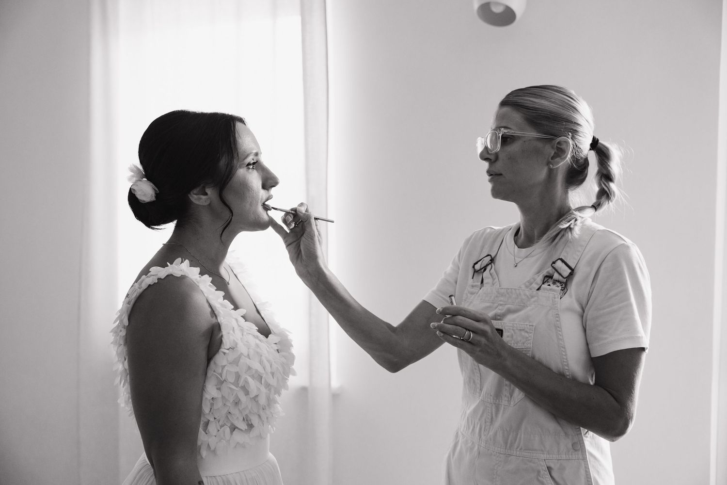 Black and white photo of a makeup artist applying lipstick to a bride in a white dress before her wedding.