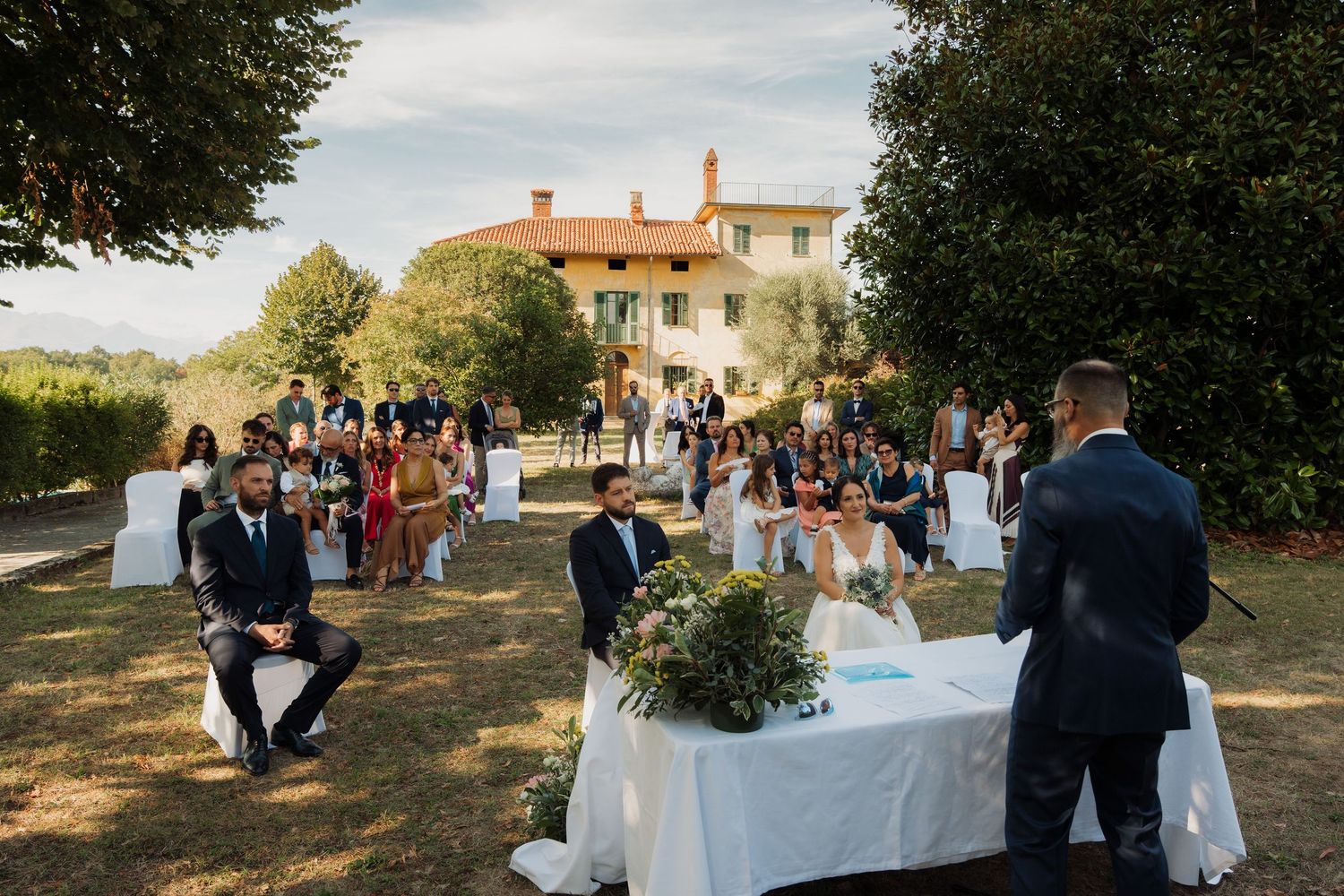 Outdoor wedding ceremony on lush green lawn with guests seated, officiant speaking, and elegant villa in background.
