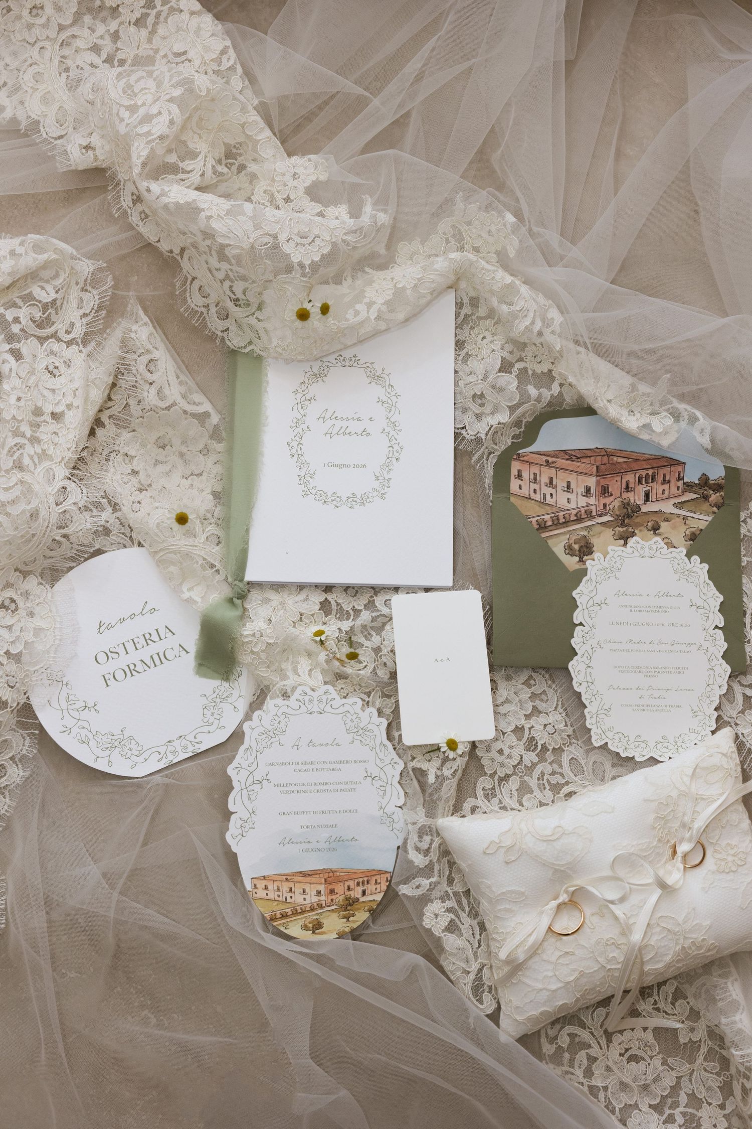 Wedding stationery flat lay with green envelope, menus, place cards, and rings on ivory lace bridal veil.
