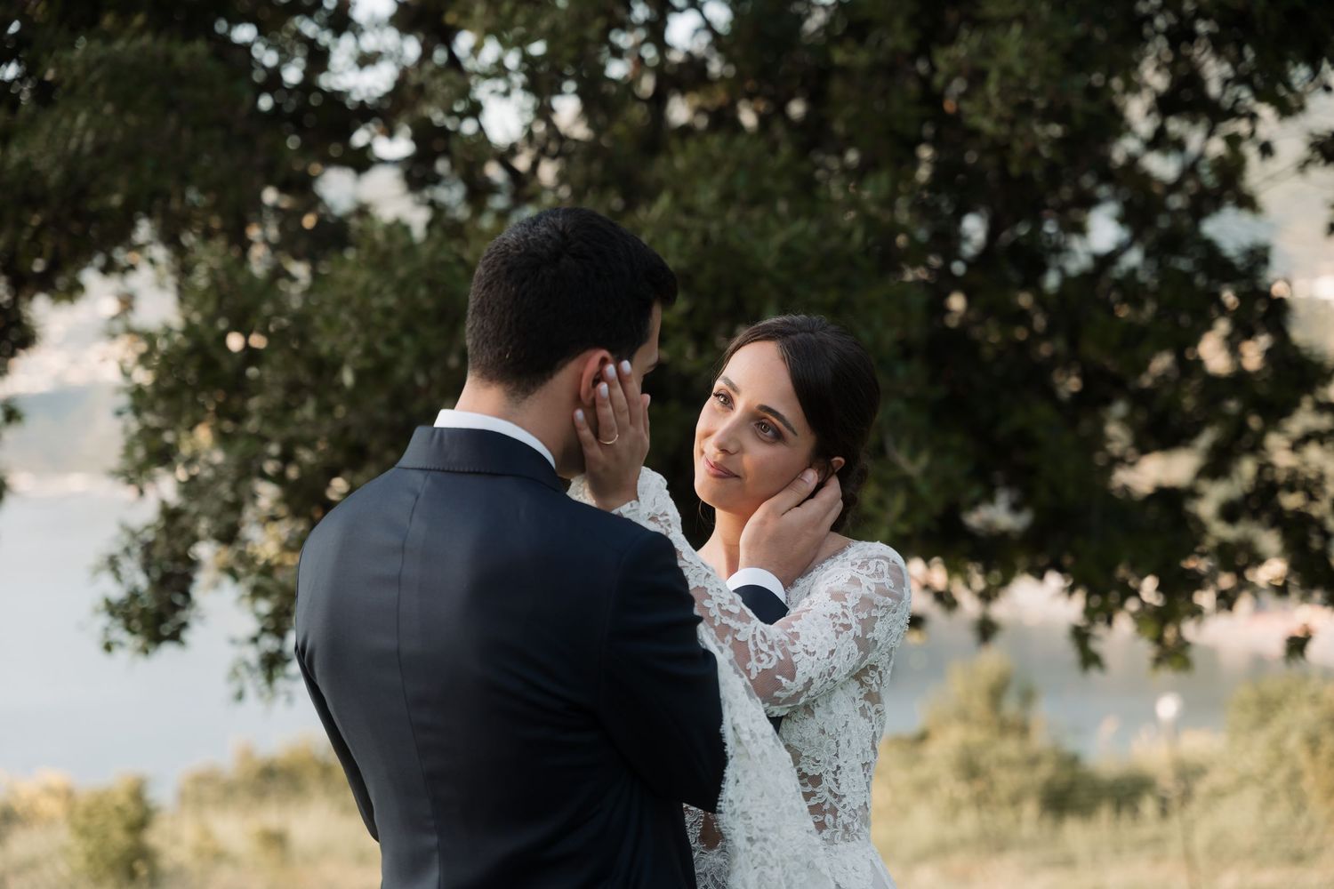 Bride tenderly touching grooms face in romantic outdoor portrait under large oak tree.