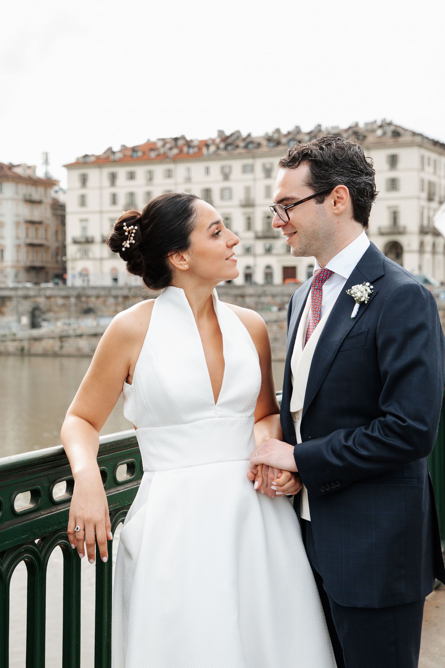 Bride and groom holding hands and gazing at each other on a bridge with city backdrop.