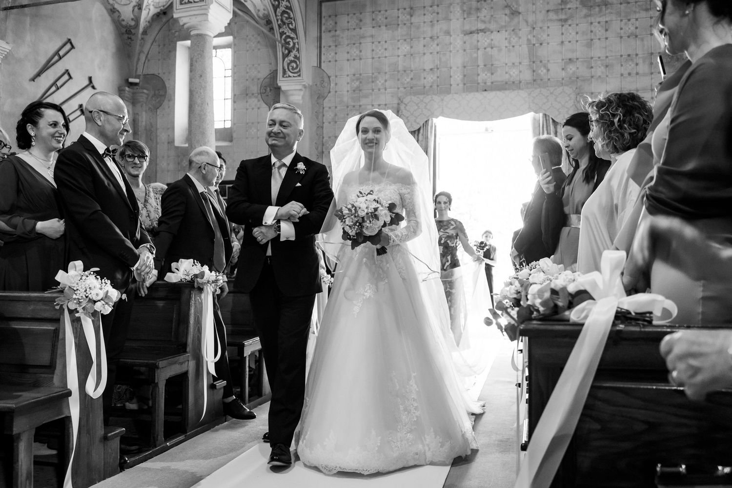 Bride walks down church aisle escorted by father in elegant black and white wedding photo.
